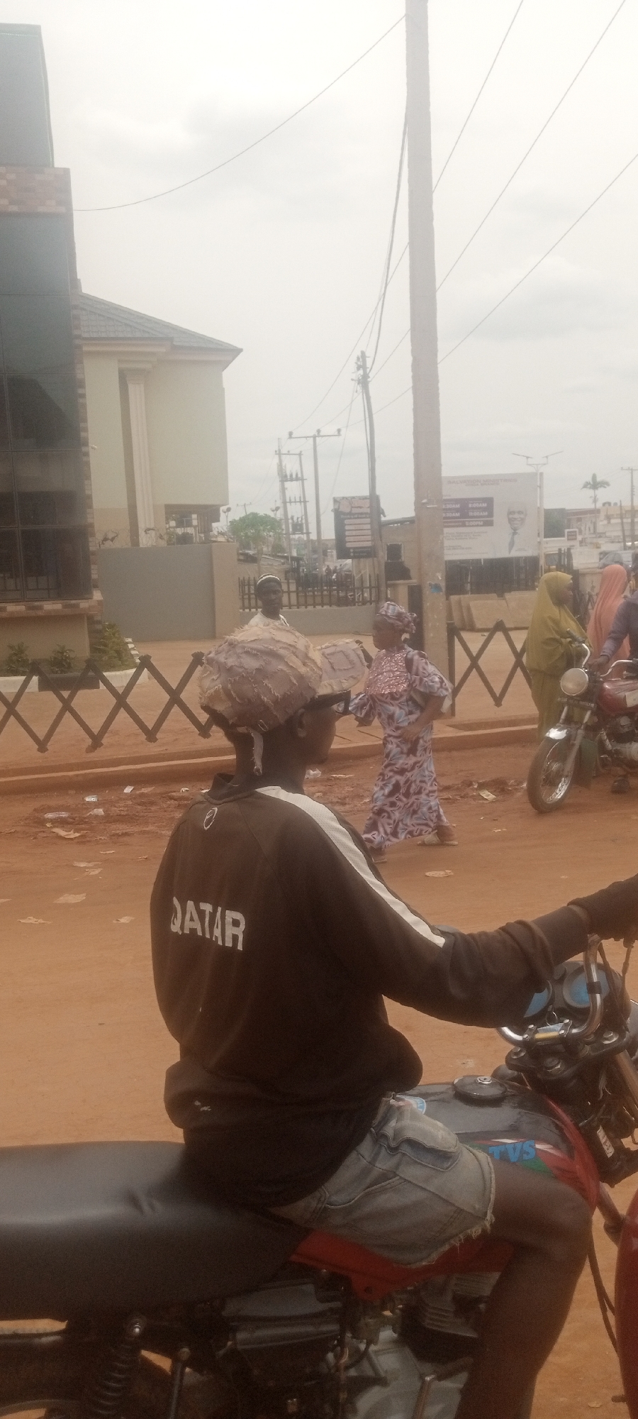 The image shows a street scene in Sabon Tasha, Nigeria. In the foreground, a person is sitting on a red and black motorcycle, wearing a dark jacket with "QATAR" printed on the back and denim shorts. They are also wearing a large, possibly woven or layered material on their head, and sunglasses.
In the background, several other individuals are visible. One person is wearing a patterned dress. Another woman is wearing a green hijab and is near a motorcycle. A billboard with a portrait and text is visible across the street behind the people.
The scene has an earthy, reddish-brown dirt road or ground, and a building and fences can be seen on the left. A tall utility pole is in the background to the right.