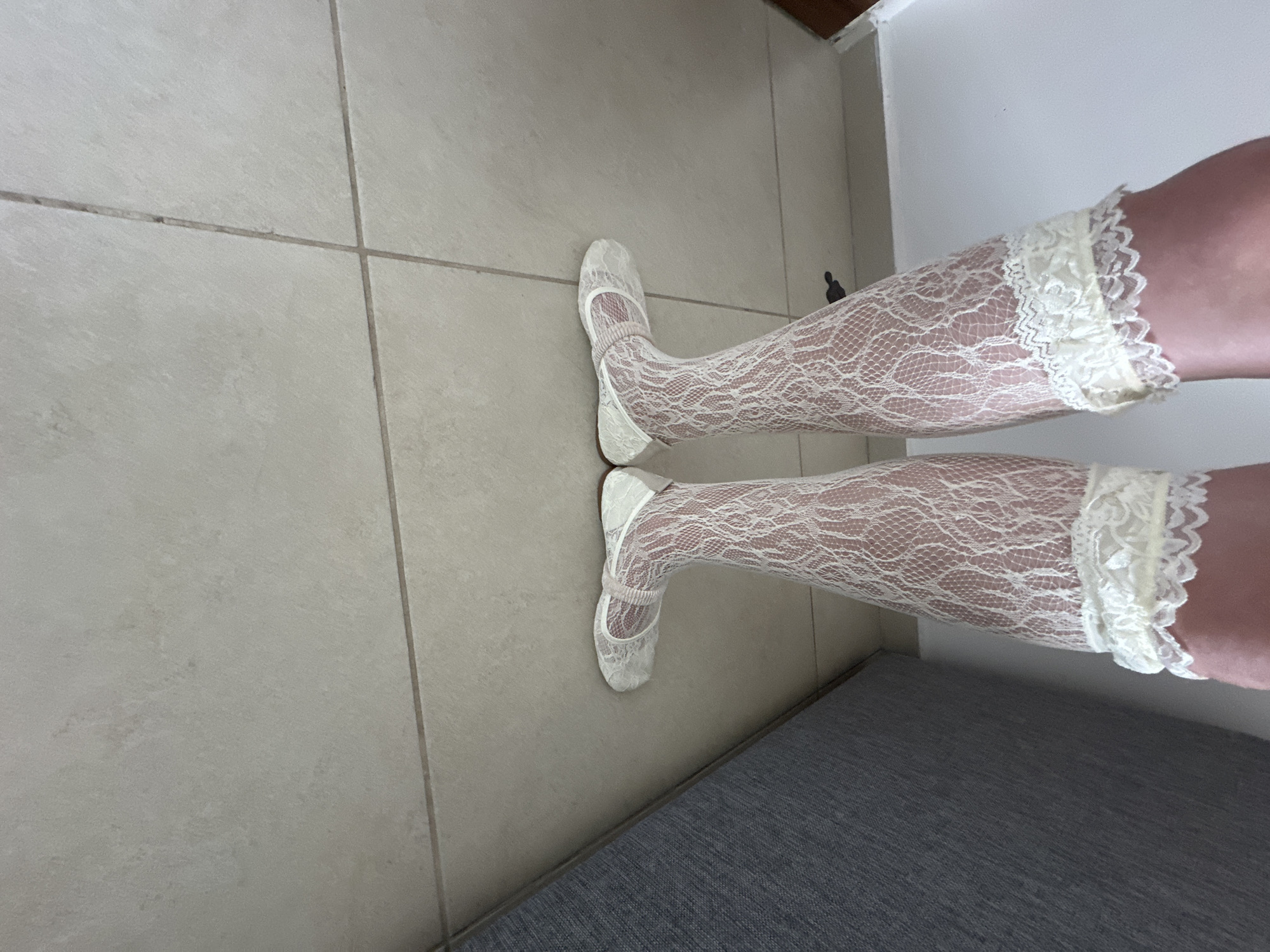 The image shows a pair of legs wearing white lace knee-high socks and matching Mary Jane-style shoes. The legs are positioned on a tiled floor with a gray upholstered object or piece of furniture visible in the lower right corner of the frame. The environment is interior, with a plain white wall visible in the upper right corner.