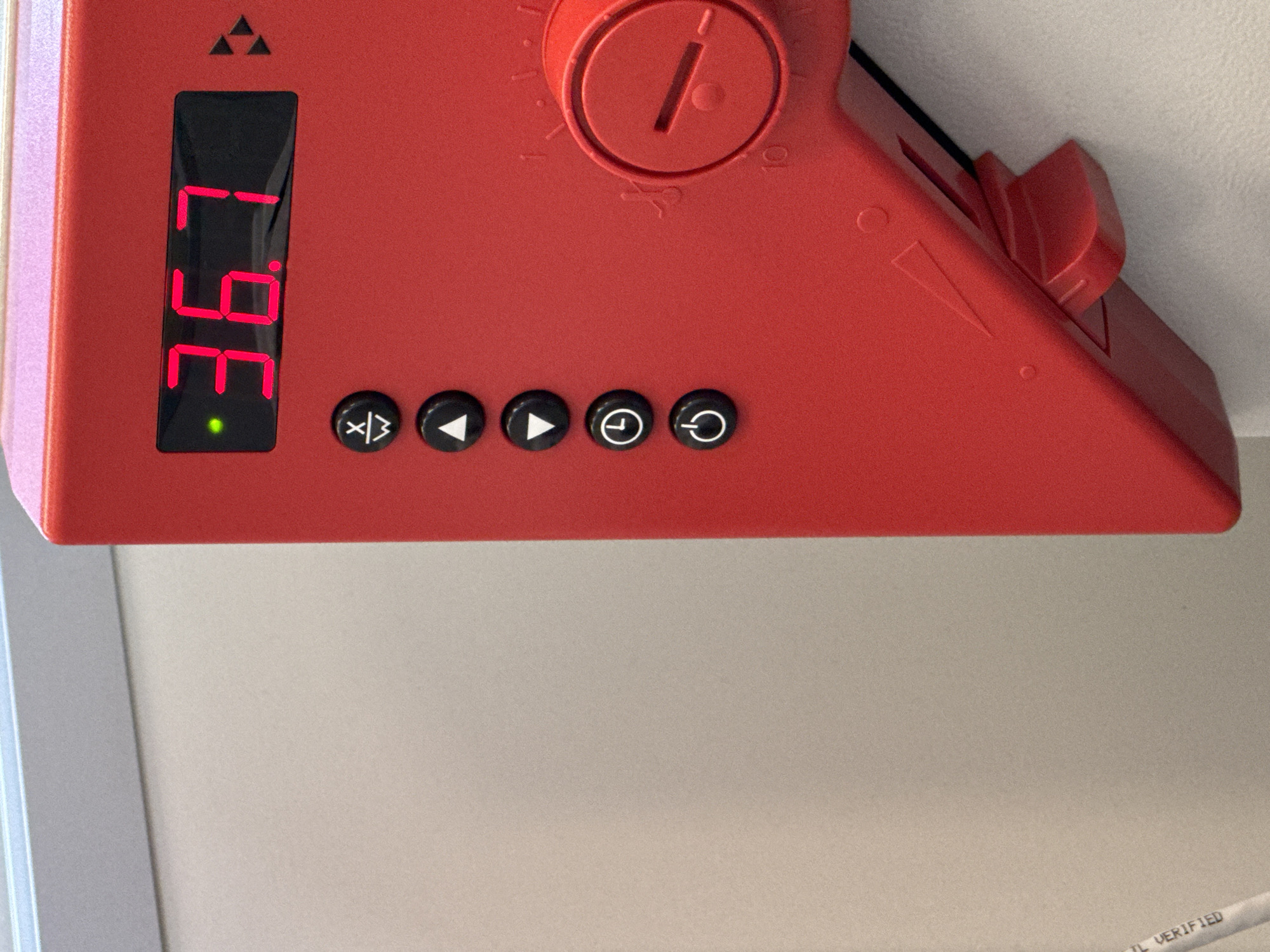 A close-up image reveals a bright red electronic device, possibly a control panel or a thermostat, mounted on a light-colored surface, likely a wall or ceiling. The device features a black vertical digital display that shows the number "36.1" in illuminated red LED segments. A small, circular green LED light glows directly below the digital readout.
To the right of the display, five black circular buttons are arranged horizontally. From left to right, their embossed white icons depict: a circle with a diagonal line and vertical bars (suggesting a mode or input selection), a left-pointing triangle (rewind or decrease), a right-pointing triangle (play or increase), a clock or timer symbol (a circle with a small line on top), and a power symbol (a circle with a vertical line).
Above the buttons, a red rotary knob with a single slot and a central dot is visible. Numerical markings around the knob, "1" and "10" being the most discernible, suggest a range of settings. The red casing also has raised triangular indicators pointing towards the right side of the device.
The background consists of a plain, light beige or off-white textured surface, with a silver-grey edge of a frame or panel visible along the left side of the image. No people are present. The illuminated display and green light indicate the device is operational. Faintly visible on a white cable in the bottom right corner of the original image are the partial words "IL VERIFIED."