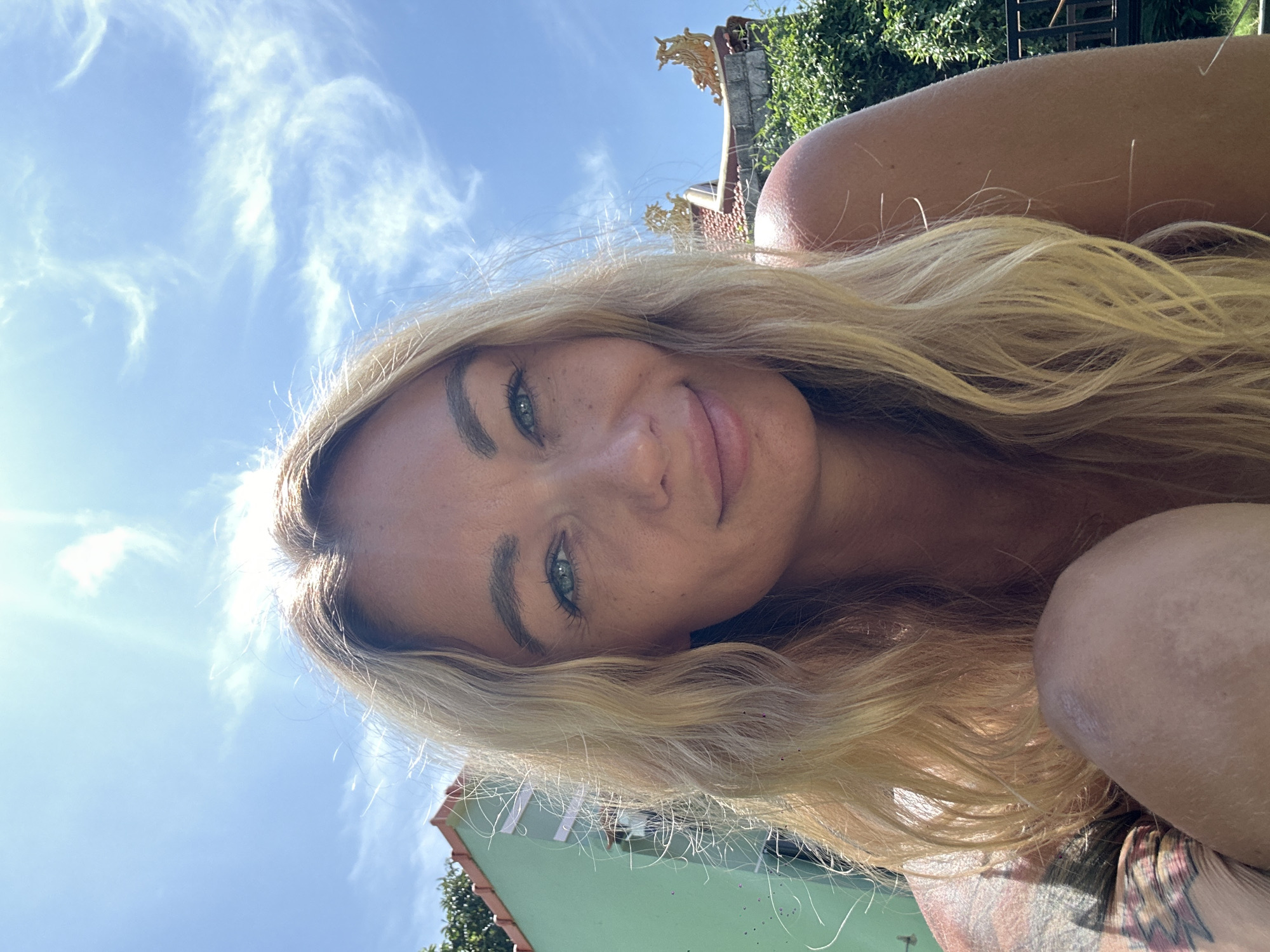 A blonde woman is pictured in a close-up, selfie-style photograph under a bright blue sky with wispy clouds. She is looking towards the camera with a slight smile. Her hair is long and wavy, framing her face. To the left of the woman, a portion of a light green building with a red-tiled roof is visible. To the right, the upper part of a building with ornate, gold-colored roof decorations and lush green foliage can be seen. The overall setting suggests a sunny outdoor location, possibly a tropical or subtropical environment. The photograph appears to be taken in Mũi Né, Vietnam.