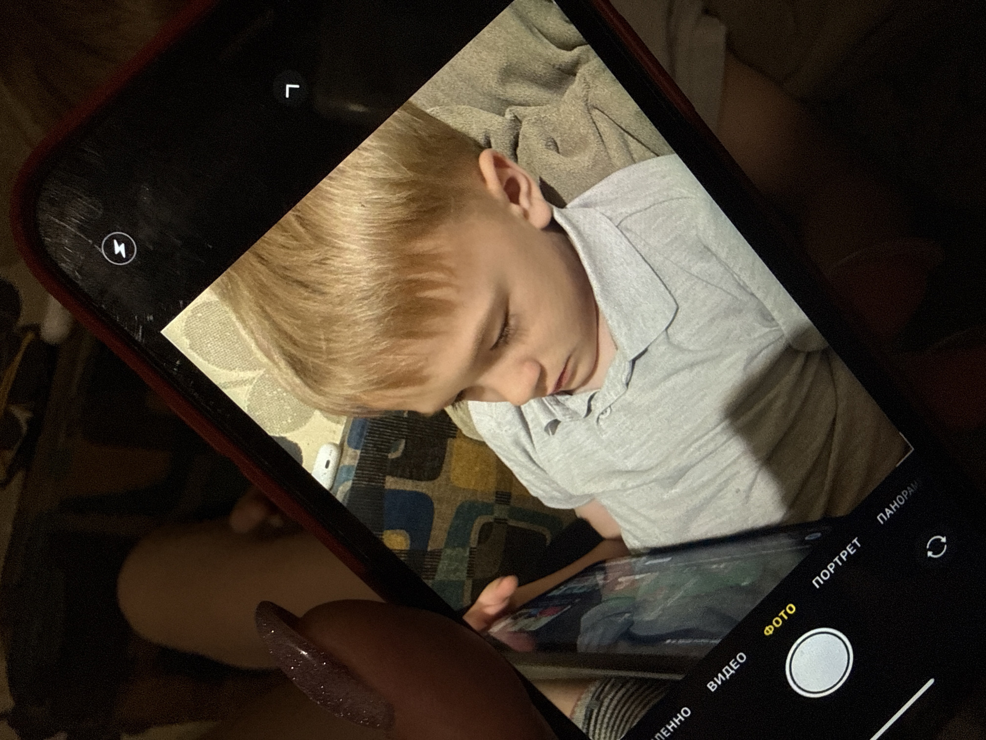 A photo shows a child sleeping, captured on a smartphone screen. The smartphone being used to take the photo has a red case and some scratches on the screen. The phone's camera interface is visible, set to "Photo" mode. The child has blonde hair and is wearing a light gray polo shirt. The child appears to be lying on a patterned surface. A person's hand with a sparkly fingernail is visible in the foreground, holding the phone. The location, Dmitrov, Russia, cannot be confirmed from the image.