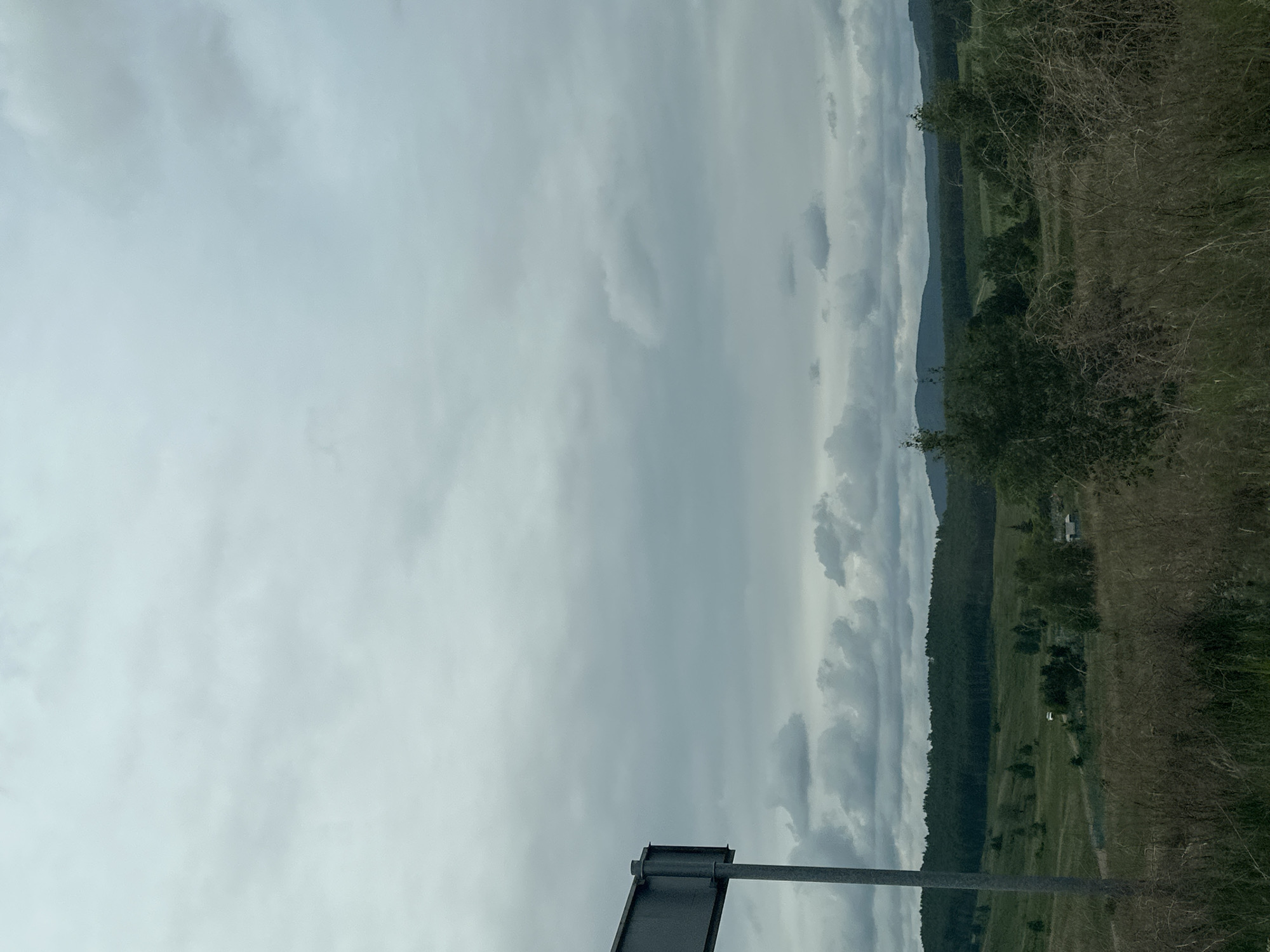The image depicts a landscape under an overcast sky. The scene includes a hillside with trees, fields, and possibly a road running through it. A road sign is visible in the foreground. The location appears to be rural.