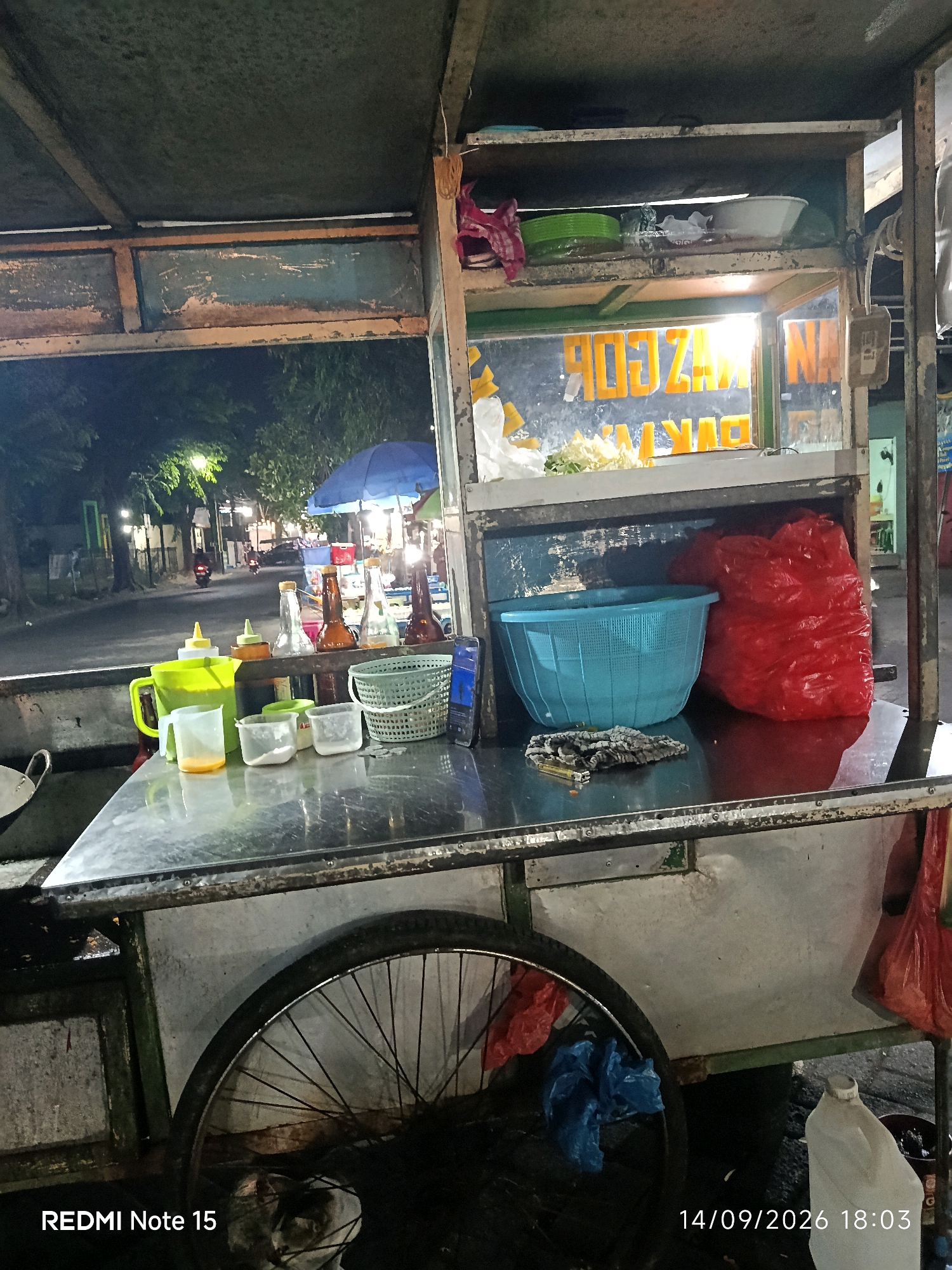 This is a nighttime, eye-level, outdoor medium shot of a food cart on a street in Keboansikep, Indonesia. The cart, made of metal with some rust and peeling paint, is loaded with various items, including bottles of condiments, plastic containers, a blue basket, and a large red plastic bag. A bicycle wheel is visible on the left side of the cart. In the background, trees, streetlights, and a few vehicles on a road are dimly lit. The scene appears to be a typical street food vendor setup at night.