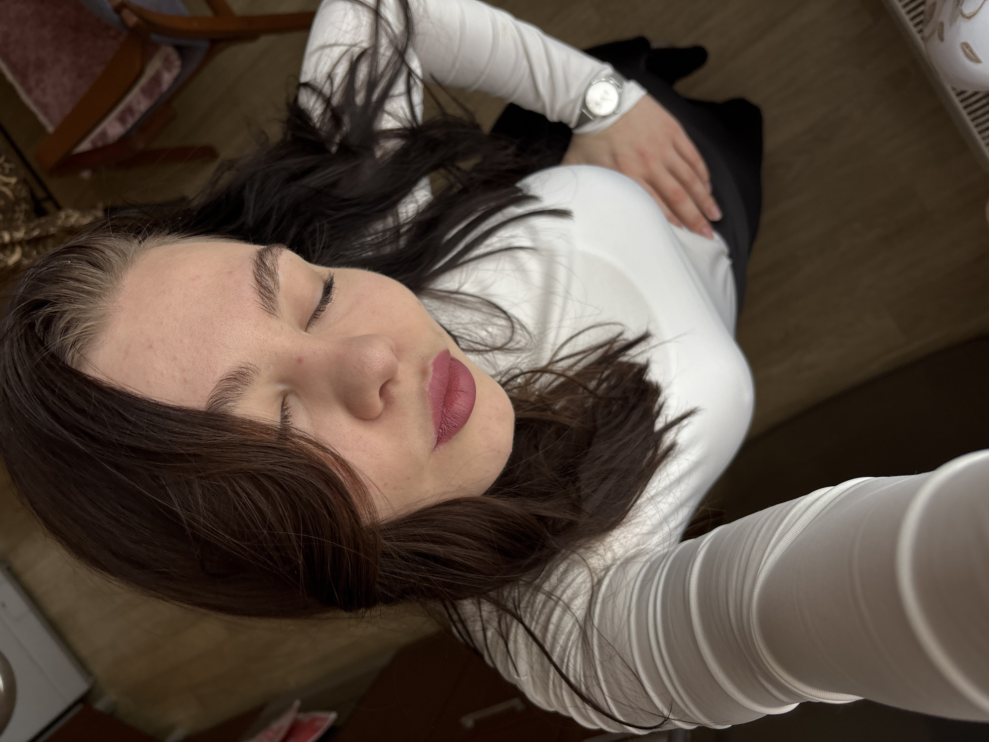 A top-down selfie captures a woman with closed eyes, full lips coated in pink lipstick, and dark brown hair, dressed in a white, long-sleeved shirt and a dark skirt or pants. A wristwatch adorns her left wrist, and her left hand rests on her waist. The background includes portions of a wooden chair and a floor, suggesting an indoor setting. It is not possible to definitively determine if the image was taken in Perm, Russia from the visual data.