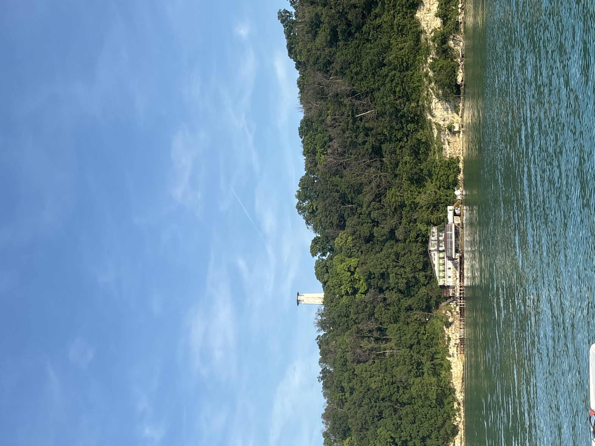 This image depicts a scenic waterside landscape under a clear blue sky, located in Unknown, Unknown. In the foreground, a tranquil body of water, possibly a lake or sea, exhibits a vibrant teal hue with numerous ripples and reflections across its surface. A small, white, indistinct object, possibly part of a boat, is partially visible at the bottom left.
The midground is dominated by a densely forested hillside rising directly from the water's edge. The hill is covered with an abundance of green trees, some appearing deciduous, and features a light-colored, rocky cliff face at its base, particularly towards the right. A multi-story building is situated at the foot of the hill, adjacent to the water. This building has a light-colored exterior with noticeable green architectural details, possibly window frames or awnings, and appears to be built on or next to a dock structure with several wooden pilings extending into the water.
Prominently positioned on the crest of the hill, above the treeline, is a slender, cylindrical white tower with a distinct, broader flat top. The sky above is a bright, clear blue, punctuated by faint, wispy white clouds and a single, thin airplane contrail streaking across the upper right quadrant. The scene is illuminated by natural daylight, casting subtle shadows. No individuals are visible in the frame.