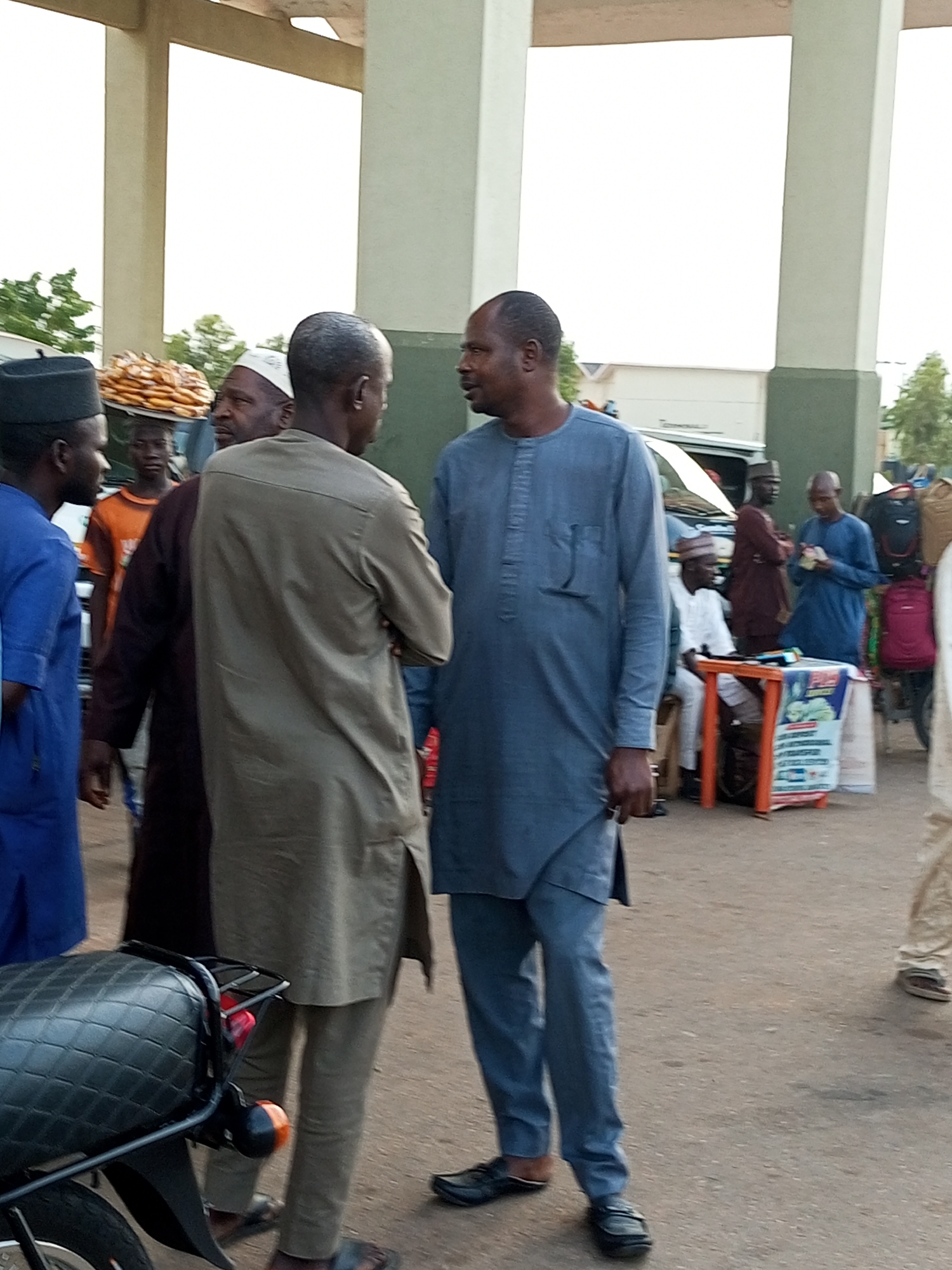The image captures a street scene in Gombe, Nigeria, likely near a marketplace or transportation hub given the presence of a motorcycle and several people engaged in conversation and transactions. Two men in traditional attire are the central focus, engaged in a discussion. The man on the left wears a light brown garment, while the man on the right is in a blue garment. Other individuals are visible in the background, including a person carrying a tray of goods on their head and others standing near a stall with merchandise. The environment appears to be outdoors, with paved ground and architectural structures suggesting a public area. The lighting indicates it is daytime, possibly late afternoon, with soft light illuminating the scene.