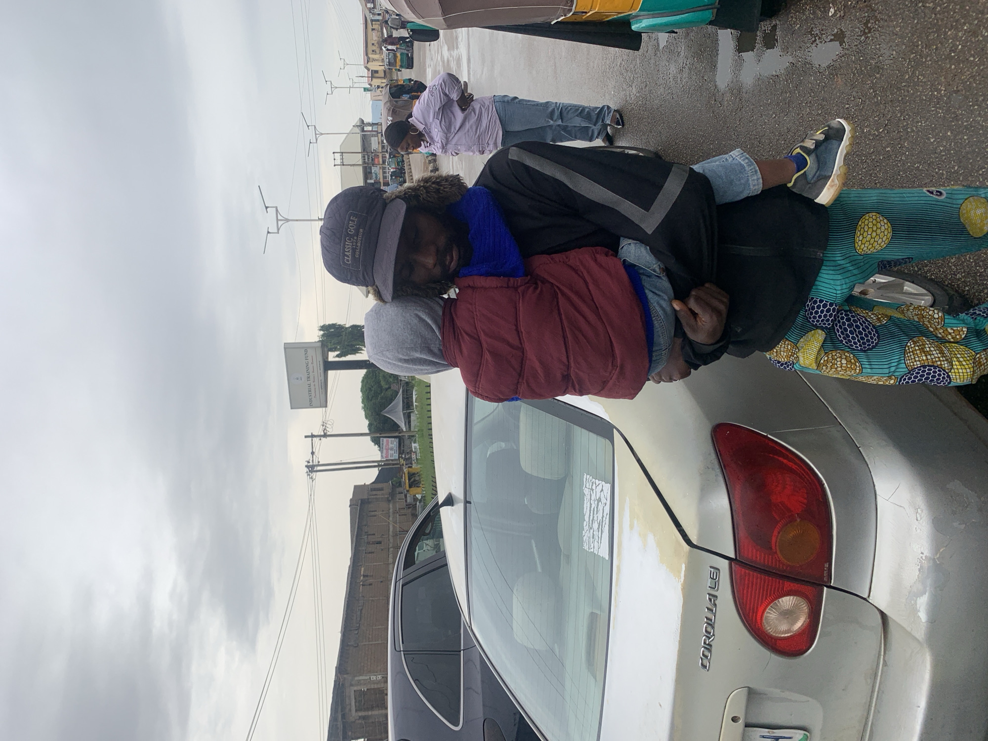 The image shows two people standing on a wet street in what appears to be an urban setting. The person in the foreground is wearing a dark jacket with a fur-lined hood, a blue scarf, and a black baseball cap with "CLASSIC GOLF" and "GOLF-GEAR" visible on it. This person is holding a child who is wrapped in a maroon vest and a grey hoodie. The child's face is partially obscured. The person in the background is wearing a light purple jacket and blue jeans. They are looking down at something on the ground.
Behind the people, a silver Toyota Corolla is parked, its rear lights and "COROLLA LEI" badging are visible. The car is a light, metallic color and appears to be slightly dirty with some yellowing paint or residue on the rear. There are other vehicles and buildings in the background, suggesting a commercial or busy area. Power lines and street lights are also visible, indicating an outdoor, public space. The ground is wet and reflective, suggesting recent rain or damp conditions. The sky is overcast and gray, indicating a cloudy day, possibly during daylight hours, but without direct sunlight. The overall mood seems to be one of transit or a brief stop on a journey. There is a sign visible in the background with some text that is not clearly legible in the provided crops, but appears to be related to training.