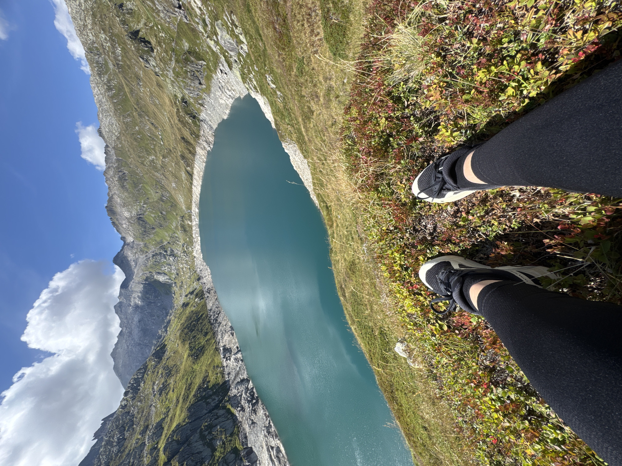 The image shows a first-person perspective of a person's legs and feet resting on a grassy hillside overlooking a large, turquoise lake. The lake is surrounded by rocky, mountainous terrain with patches of green grass and scrub. The sky is blue with scattered white clouds. The person is wearing black leggings and athletic shoes with white soles and black laces. The vegetation in the foreground consists of low-lying bushes with red and green leaves, and dry grass. The overall setting is a scenic mountain landscape. The location is the Passo del San Gottardo in Airolo, Switzerland.