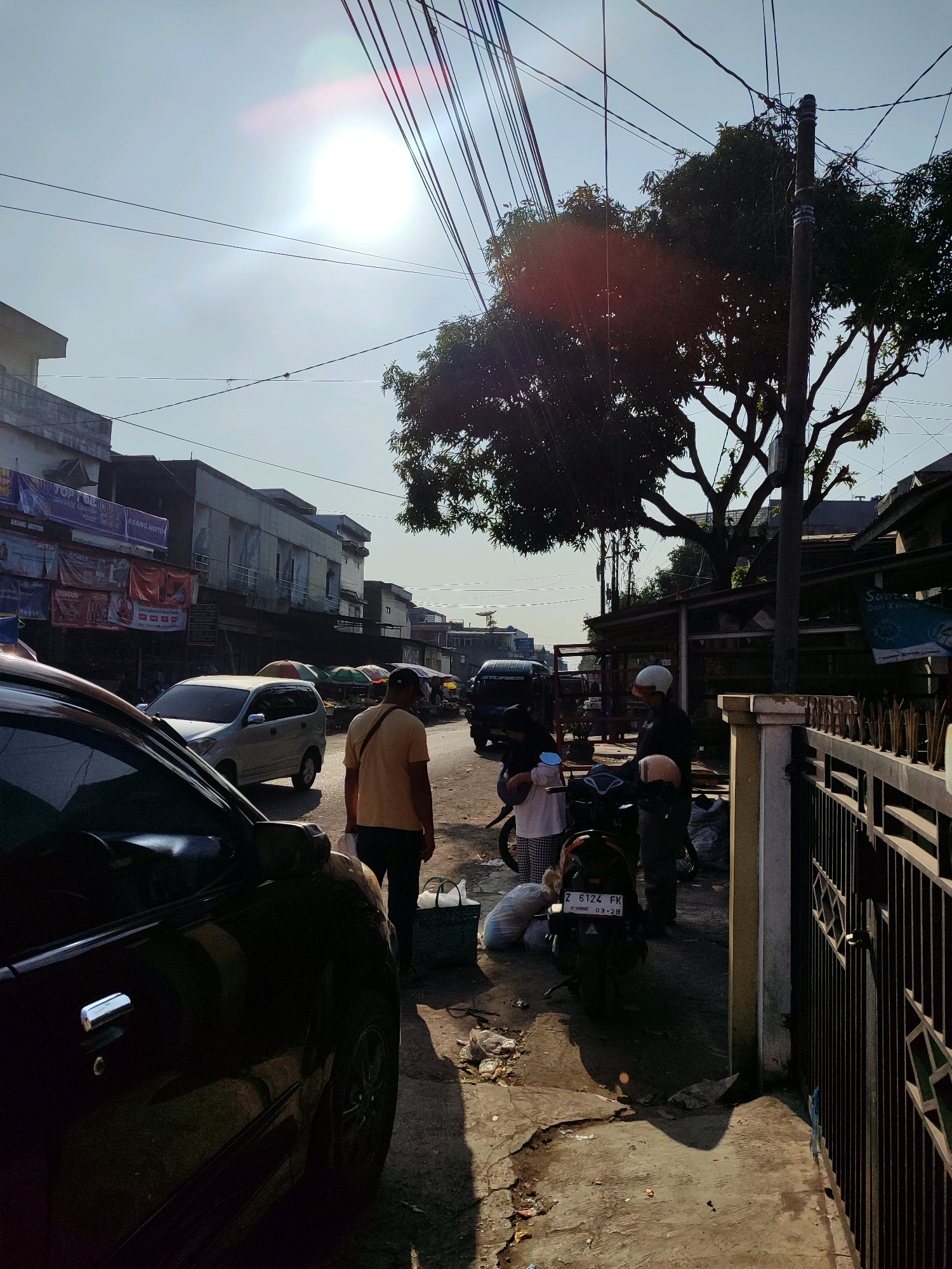 The image depicts a street scene in Garut, Indonesia, under a bright, hazy sun. A black car is parked on the left side of the frame, with its side and wheel visible. Further down the street, a light-colored minivan is driving. On the right side of the road, a man in a yellow shirt stands, holding a plastic bag. Behind him, a motorcycle is parked, and two people, one wearing a white shirt and the other a helmet, are near it. A black metal fence runs along the right edge of the image, bordering a building. The street is lined with buildings and trees, and there are various market stalls with umbrellas set up along the side of the road. Power lines crisscross the sky. The ground is paved and appears somewhat worn, with scattered debris. The overall impression is that of a bustling street, possibly a market area, during the day.