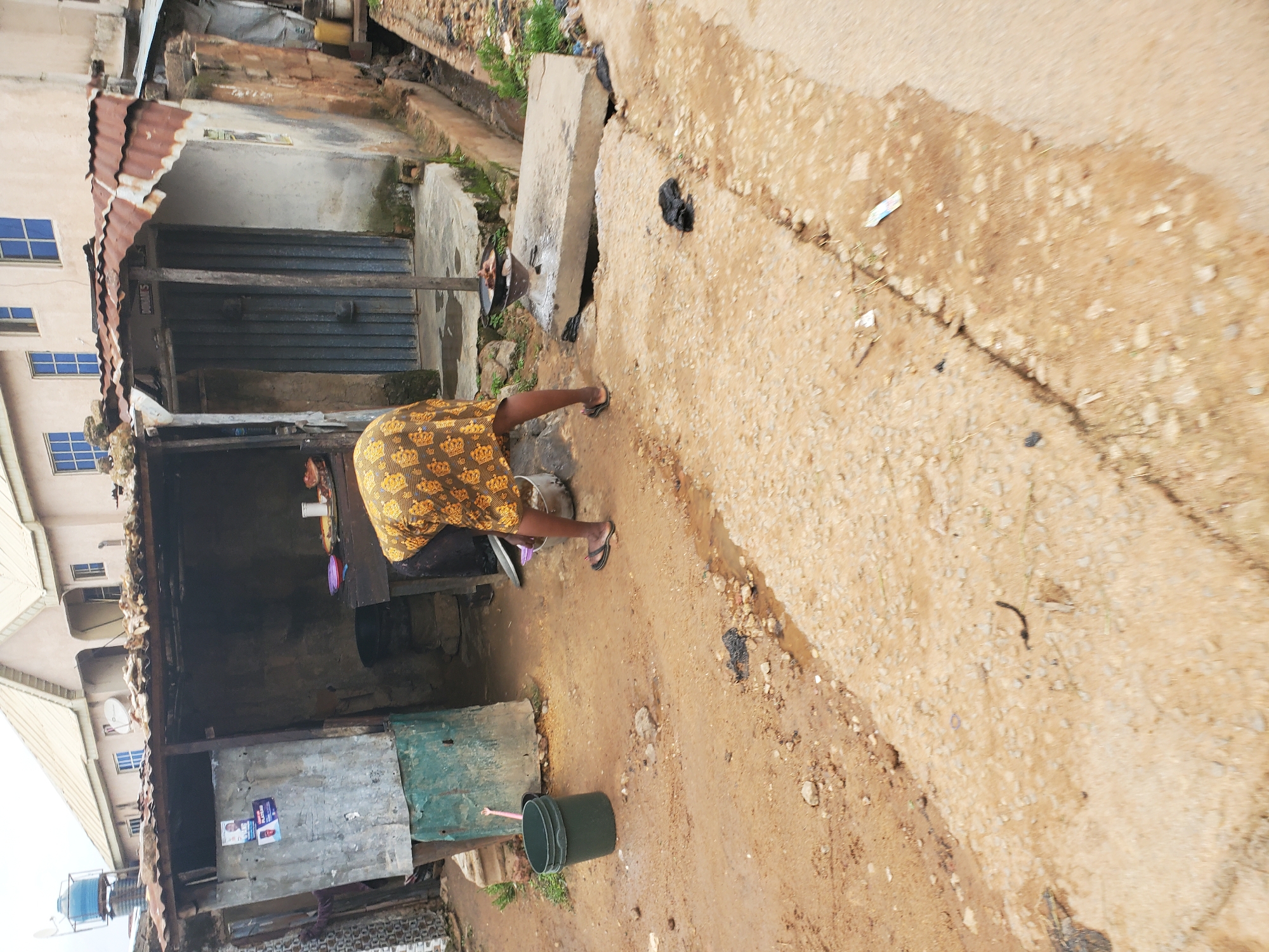 A person wearing a yellow and brown patterned dress is bending over, sweeping the ground with a broom in front of a ramshackle outdoor stall. The stall, constructed from corrugated metal and wood, appears to be a food vendor's stand, with cooking utensils and food visible on a counter inside. A large green bucket and a cooking pot are on the ground outside the stall. The ground is dirt and gravel, with some puddles of water, suggesting recent rain. In the background, a multi-story building with beige walls and blue-framed windows stands under an overcast sky. The scene appears to be in a densely populated urban area, possibly a market or street side, consistent with the city of Jos, Nigeria.