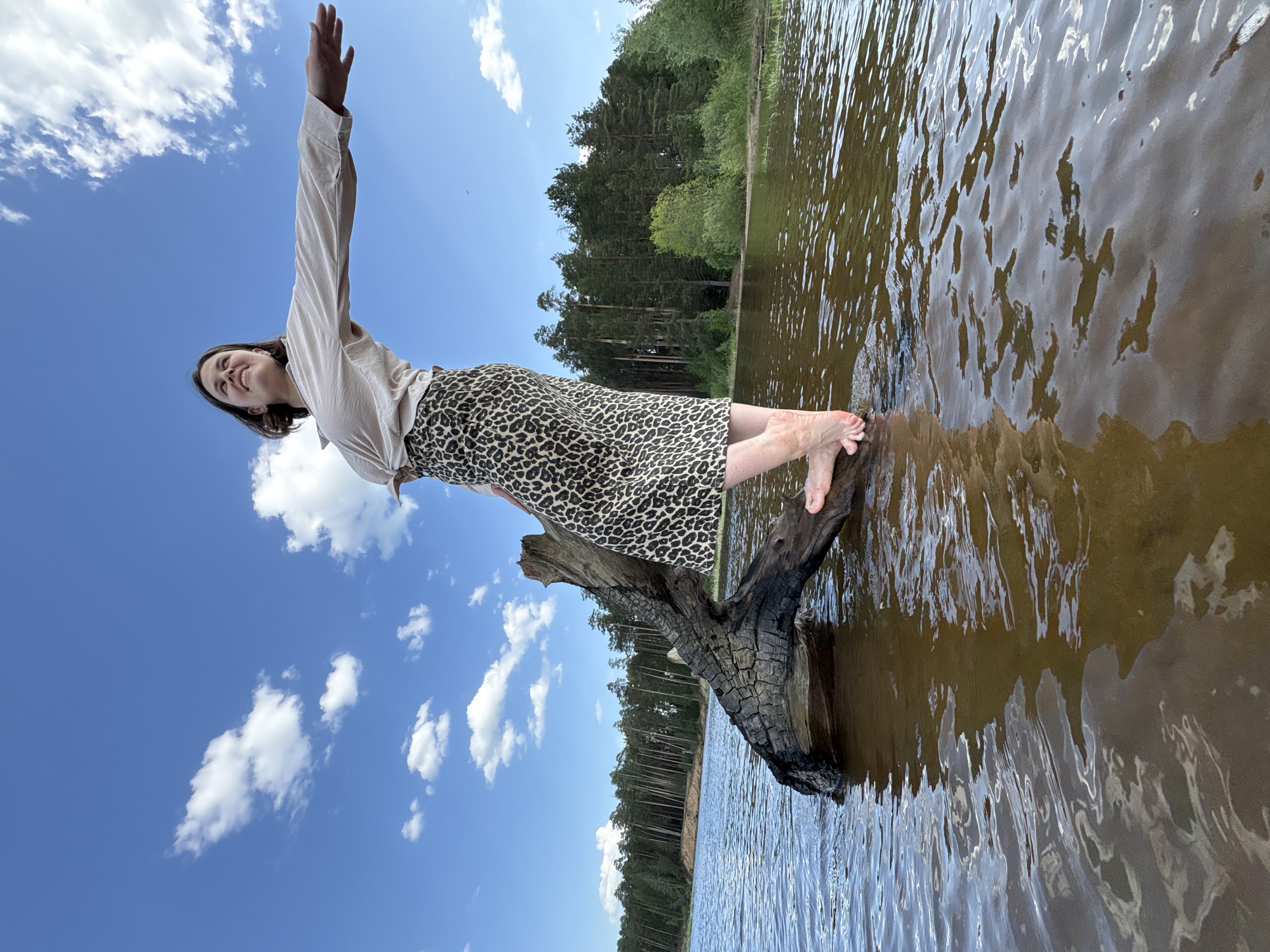 A woman stands on a partially submerged burnt log in a lake. She is wearing a light-colored blouse and a leopard-print skirt. Her right arm is raised in the air. The lake reflects the surrounding forest. The sky is blue with scattered clouds. This photo was taken near the city of Chaikovsky in the Perm Krai region of Russia.