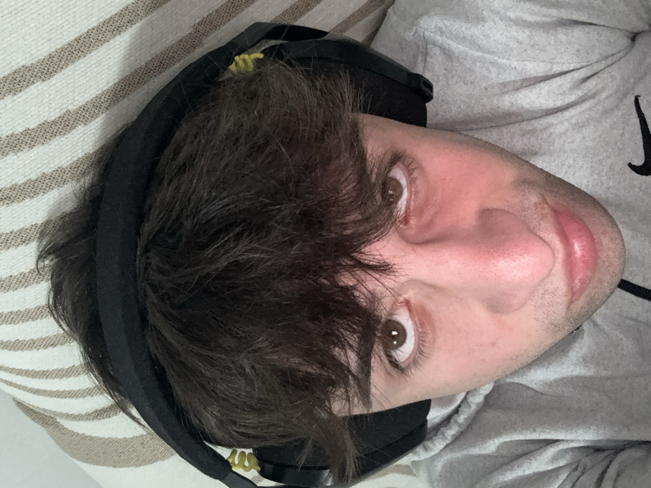 The image captures a young male subject indoors, viewed from a slightly low angle. He has dark, disheveled hair, brown eyes, and a neutral expression, looking directly into the camera. He wears a light grey hooded sweatshirt with a black Nike swoosh logo visible on the lower right chest, and black over-ear headphones with yellow accents on the cabling. The background is a striped fabric, likely a cushion or textile surface, featuring alternating light beige and off-white horizontal stripes. No specific geographic identifiers are present in the image to confirm a location in Selanovtsi, Bulgaria.