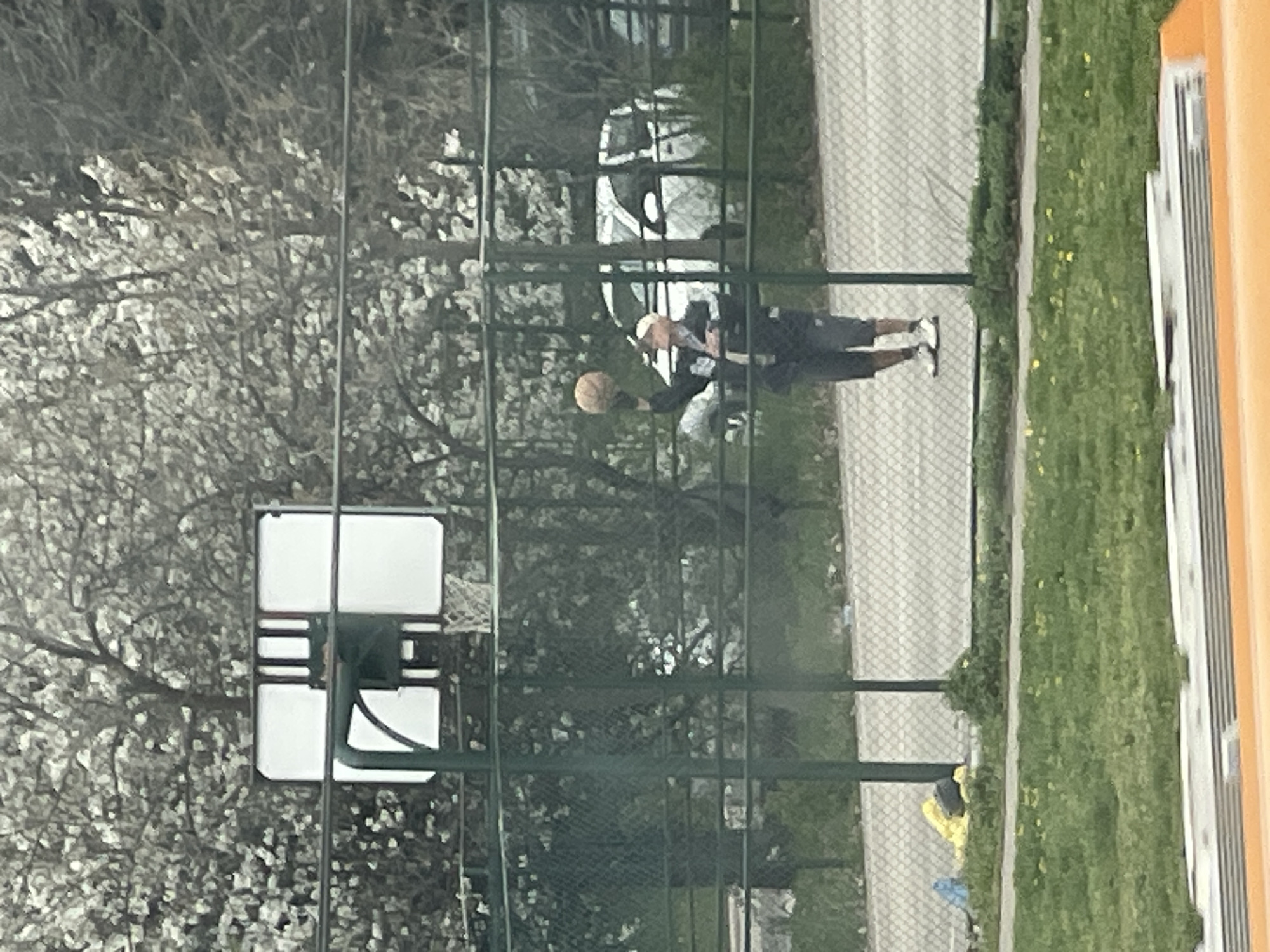 The image shows a basketball court in Sofia, Bulgaria, with a basketball hoop and net positioned on the left. A man, wearing dark shorts and a light-colored top, is holding a basketball. He is standing on the court's surface, which is surrounded by a green fence. In the background, there is a street, trees, and a vehicle. The image seems to have been taken from an elevated position, possibly from a building.