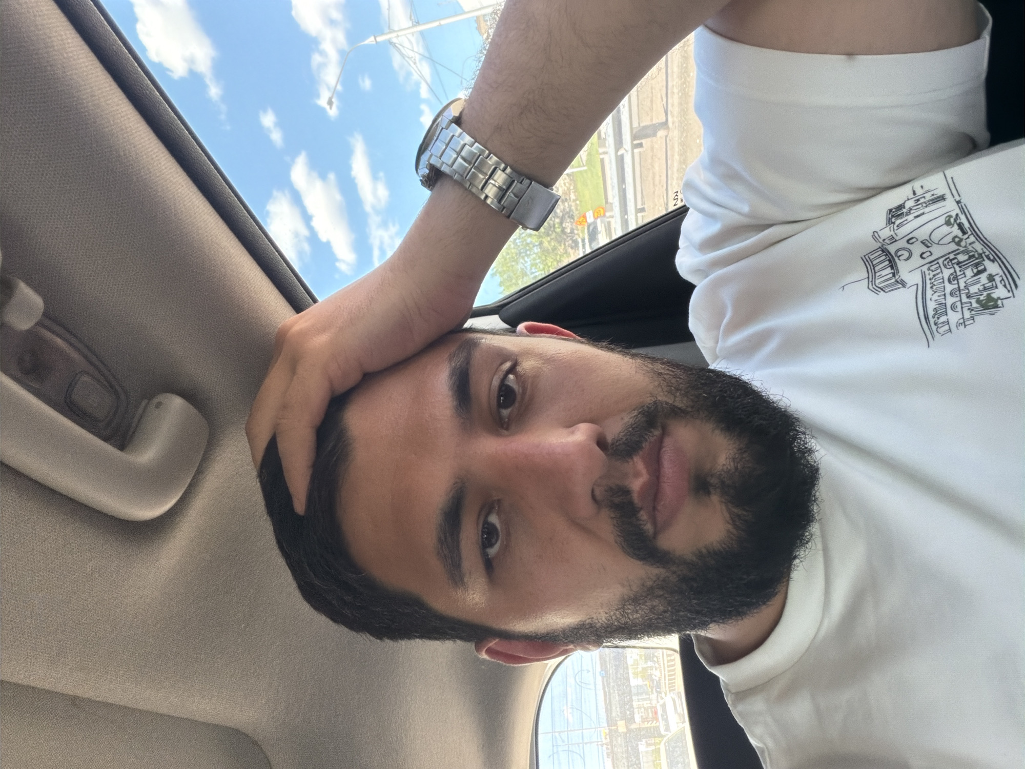 A medium shot of a man with dark hair and a full, dark beard in the driver's seat of a car, shot from a slightly low angle. The man is fair-skinned and is wearing a white t-shirt with a printed design. A metal wristwatch is visible on his left wrist. His left hand is placed on his head. The interior of the car is light gray. Through the car window, the city of Krasnoyarsk, Russia is visible.