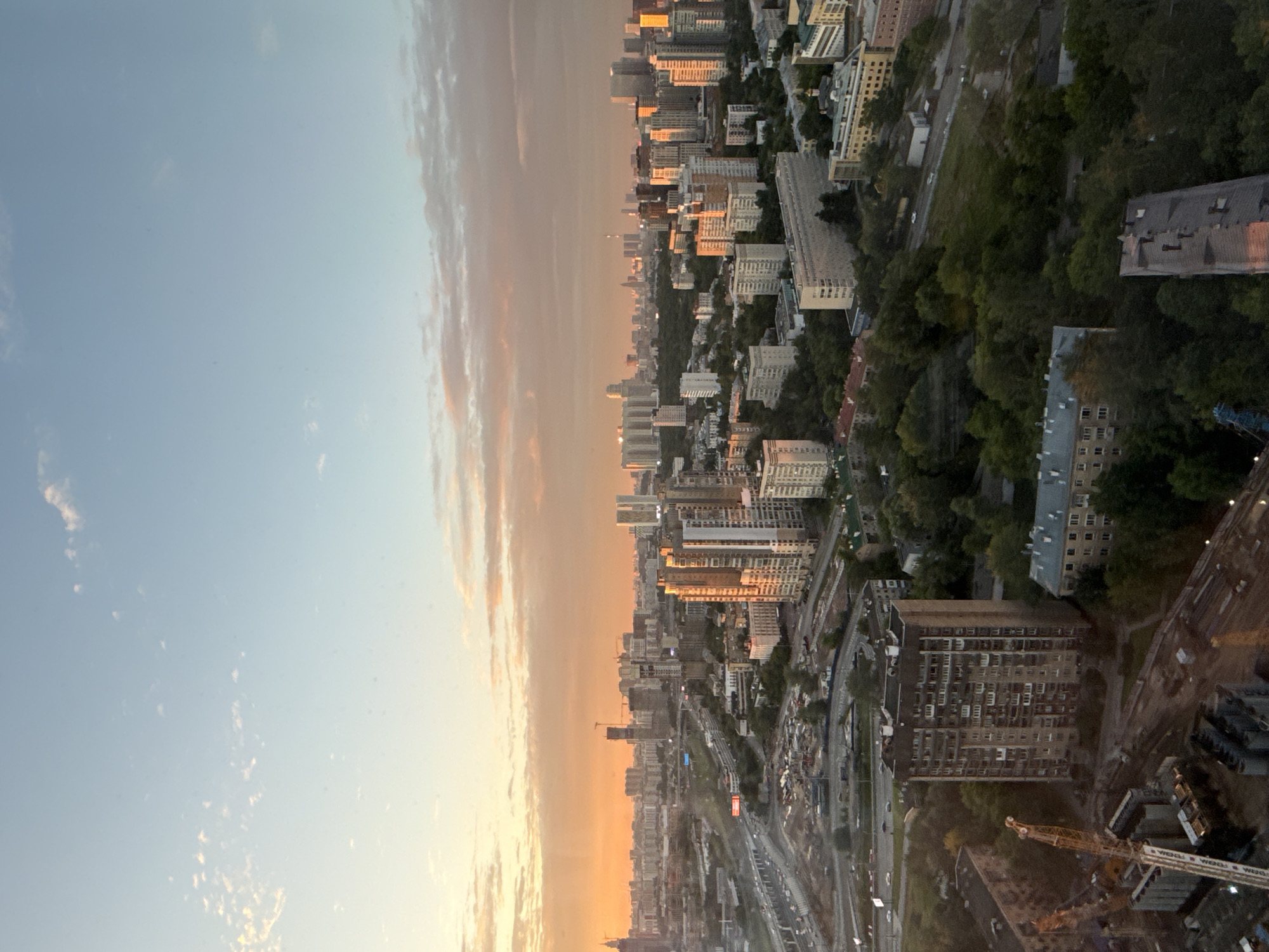 This is an aerial, wide-angle photograph of a city skyline at dusk. The sky transitions from a pale blue at the top to a vibrant orange and pink hue near the horizon, indicating the setting sun. Clouds are scattered across the sky, reflecting the warm light.
The scene is dominated by a dense urban landscape with numerous buildings of varying heights and architectural styles. In the foreground, several tall, multi-story residential buildings are visible. One particularly prominent building in the lower-left quadrant is dark-colored with many balconies. Adjacent to it, construction cranes are visible, suggesting ongoing development.
Further into the mid-ground, more residential buildings and some taller, modern skyscrapers are present. A multi-lane highway or road system curves through the city, with a few cars visible. Patches of green trees and parkland are interspersed between the buildings, particularly in the mid-ground and background.
In the distance, a sprawling cityscape extends towards the horizon, with many more buildings forming a dense urban fabric. The warm light of the sunset casts long shadows and highlights the facades of some of the buildings. The overall impression is that of a large, active metropolitan area during the evening hours. This scene depicts an area within the city of Khimki, Russia, as evidenced by the distinct architectural styles and urban planning visible.