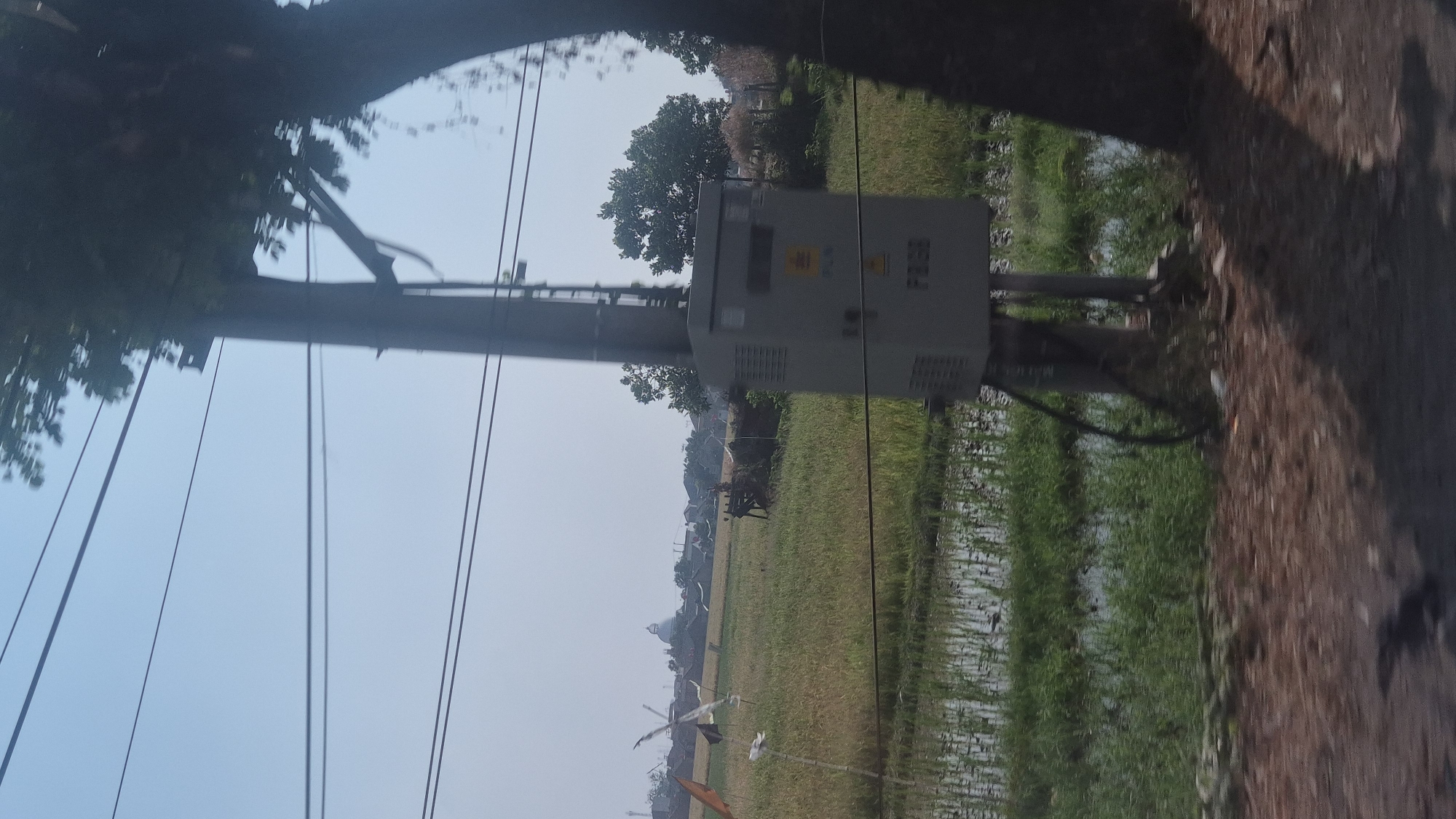 The image depicts a view from a vehicle, likely a car, traveling along a road in Karawang, Indonesia. The scene is characterized by a rural landscape. In the foreground, a dirt road is visible, partially covered by shadows from a large tree to the right. The middle ground features a rice paddy, with young rice plants growing in flooded fields and some dry, harvested areas. Scattered across the paddy are several colorful flags and some simple wooden structures.
To the right of the paddy, a utility pole stands tall, supporting overhead power lines that stretch across the frame. Attached to the pole is a gray electrical box with the "PLN" logo and the designation "F2GB" visible on it. A thick black cable runs down from the box along the pole. Further in the background, a cluster of buildings, including a prominent domed structure, suggests a village or small town. The sky is clear and blue. The overall impression is of a roadside view of agricultural land and infrastructure in a semi-rural Indonesian setting.