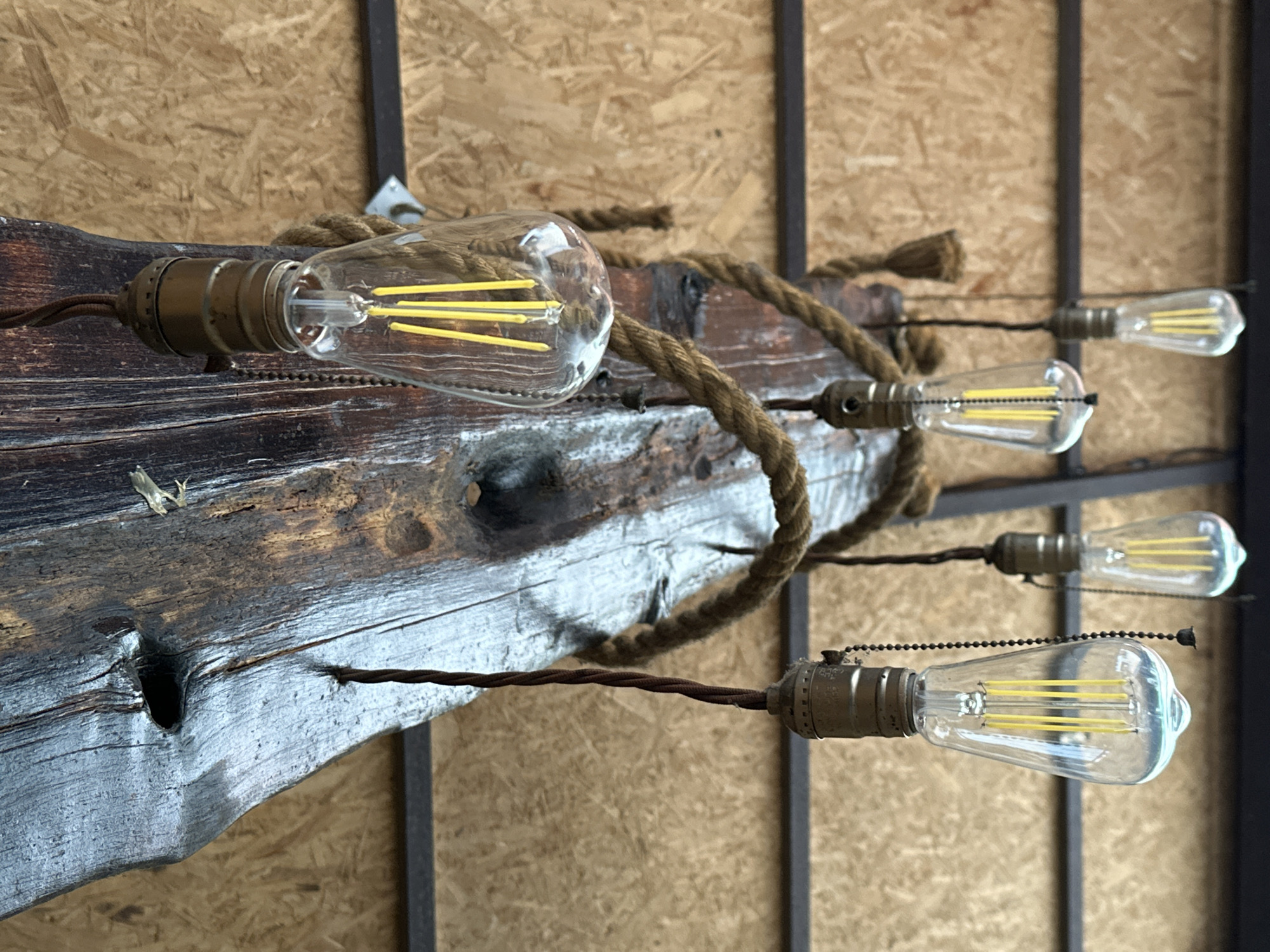 This media file showcases a rustic light fixture with a vintage aesthetic, mounted against a textured, light-brown wooden plank wall. The fixture consists of a weathered, dark brown wooden beam serving as the base, from which multiple Edison-style light bulbs are suspended. Each bulb is encased in clear glass, revealing intricate filament designs in a bright yellow hue. The bulbs are connected by dark, twisted electrical wires, some of which are partially obscured by a thick, braided rope that is also wrapped around the wooden beam.
The lighting appears to be artificial, as suggested by the illuminated bulbs. The overall impression is one of a dimly lit, possibly indoor setting, with a focus on handcrafted or repurposed materials. The wooden beam has a worn, aged appearance, with visible grain patterns and some rough textures, suggesting it may be reclaimed wood. The rope adds to the rustic, industrial, or nautical theme. The background, a subtly patterned wooden wall, is out of focus, directing the viewer's attention to the intricate details of the light fixture.
There are no people visible in the image. The arrangement of the bulbs, suspended at different heights and distances from the main beam, creates a visually dynamic composition. The light cast by the bulbs is warm and inviting, contributing to a cozy atmosphere. No text is visible in the media file.