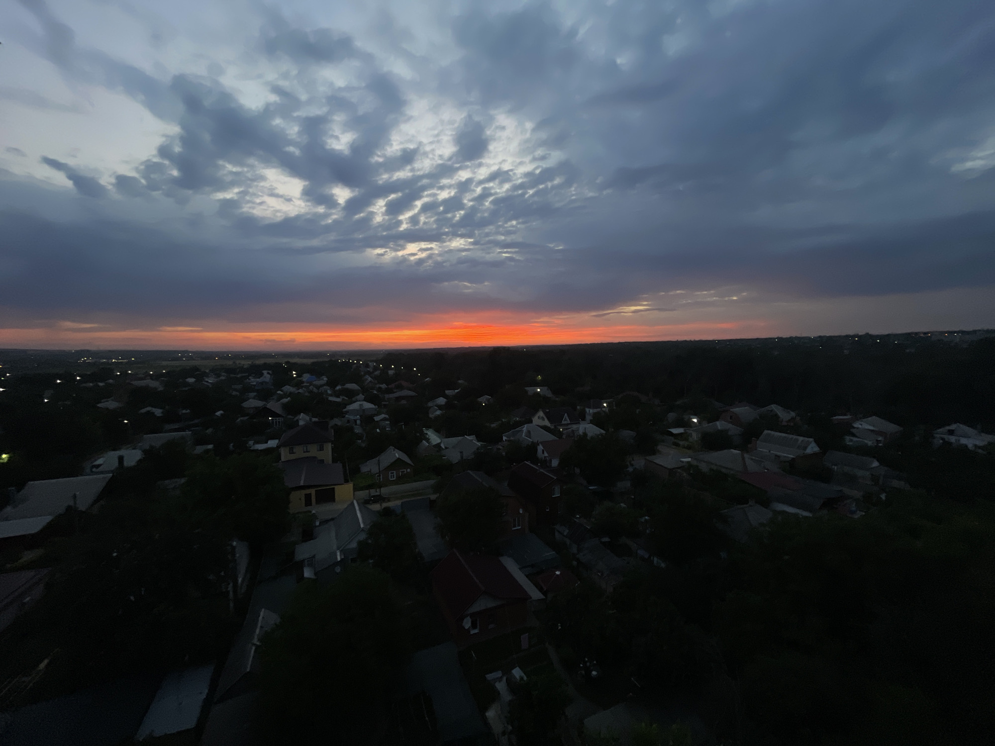 An aerial view captures a residential area in Novocherkassk, Russia, at dusk. The sky is filled with dramatic clouds, transitioning from a deep gray at the top to a vibrant orange-red glow near the horizon, indicating the setting sun. Below the sky, a dense collection of houses with varied rooflines fills the landscape, interspersed with trees. Many of the houses have visible roofs, some appearing to be made of metal or tiles, and a few lights are illuminated, suggesting inhabitants are present. The overall scene is peaceful, with the fading daylight casting long shadows over the rooftops and trees, hinting at the transition from day to night.