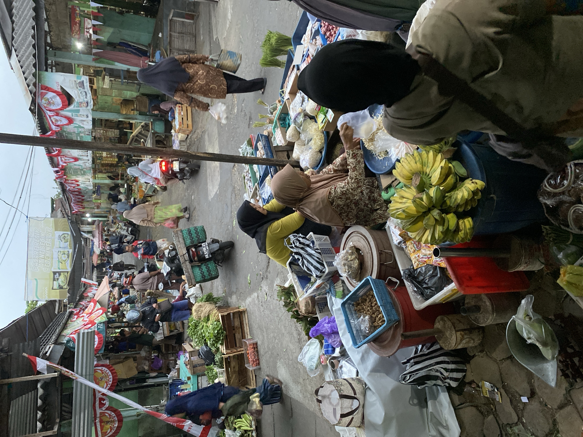 The image depicts a bustling outdoor market scene in Ambarketawang, Indonesia. Numerous vendors and shoppers are present, many of whom are wearing hijabs and traditional Indonesian attire. The market stalls are filled with a variety of fresh produce, including leafy green vegetables, bananas, and what appear to be onions or shallots. Several motorcycles are parked or being ridden through the market, indicating a common mode of transportation. Red and white Indonesian flags are prominently displayed, suggesting a festive occasion or national holiday. The architecture of the market stalls features corrugated metal roofs and open-air structures, typical of traditional Indonesian markets. The ground is paved, and there is a general sense of activity and commerce. The lighting suggests it is either early morning or late afternoon, with an overcast sky.