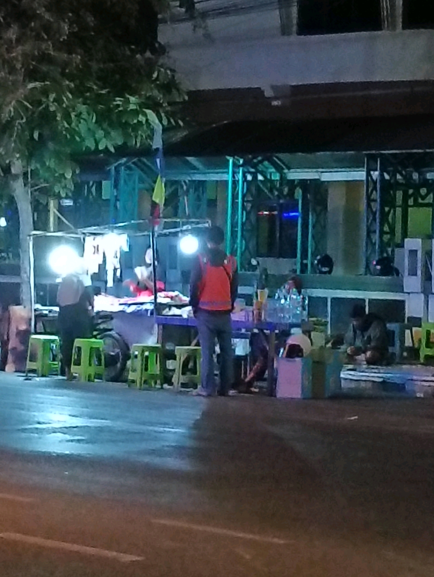 The image captures a nighttime scene on a street in Purworejo, Indonesia, likely near a food stall or market. In the foreground, a wet street reflects the ambient lighting. Several bright green plastic stools are arranged on the sidewalk. A food stall is set up with tables, displays of goods, and bright overhead lights illuminating the area. Two individuals are visible in front of the stall. One, wearing a red vest and dark pants, stands facing away from the camera. Another person, partially obscured, stands to the left of the stall. In the background, there are more stalls and structures, possibly part of a commercial area, with some glowing blue lights visible. The overall atmosphere suggests a lively, albeit somewhat hazy, street scene after dark.