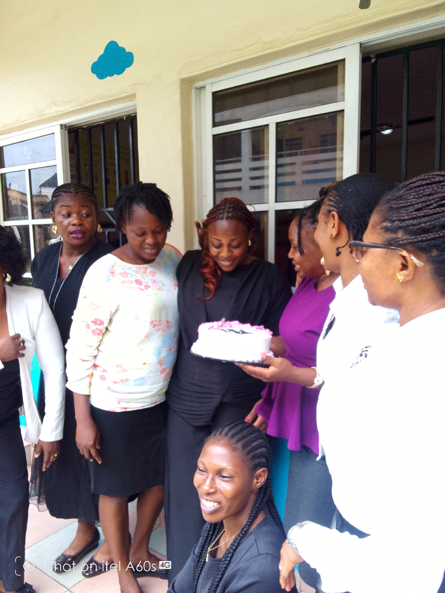 The image depicts a group of approximately eight women gathered outdoors. One woman holds a cake adorned with pink frosting. The women appear to be observing the cake with varying expressions. They are dressed in a mix of casual and semi-formal attire, including dresses, blouses, and skirts in various colors such as black, purple, white, and floral patterns. The background features a building with windows and a painted blue cloud-shaped design on the wall. The image includes the words "Shot on itel A60s 4G," suggesting the location is Idimu, Nigeria.