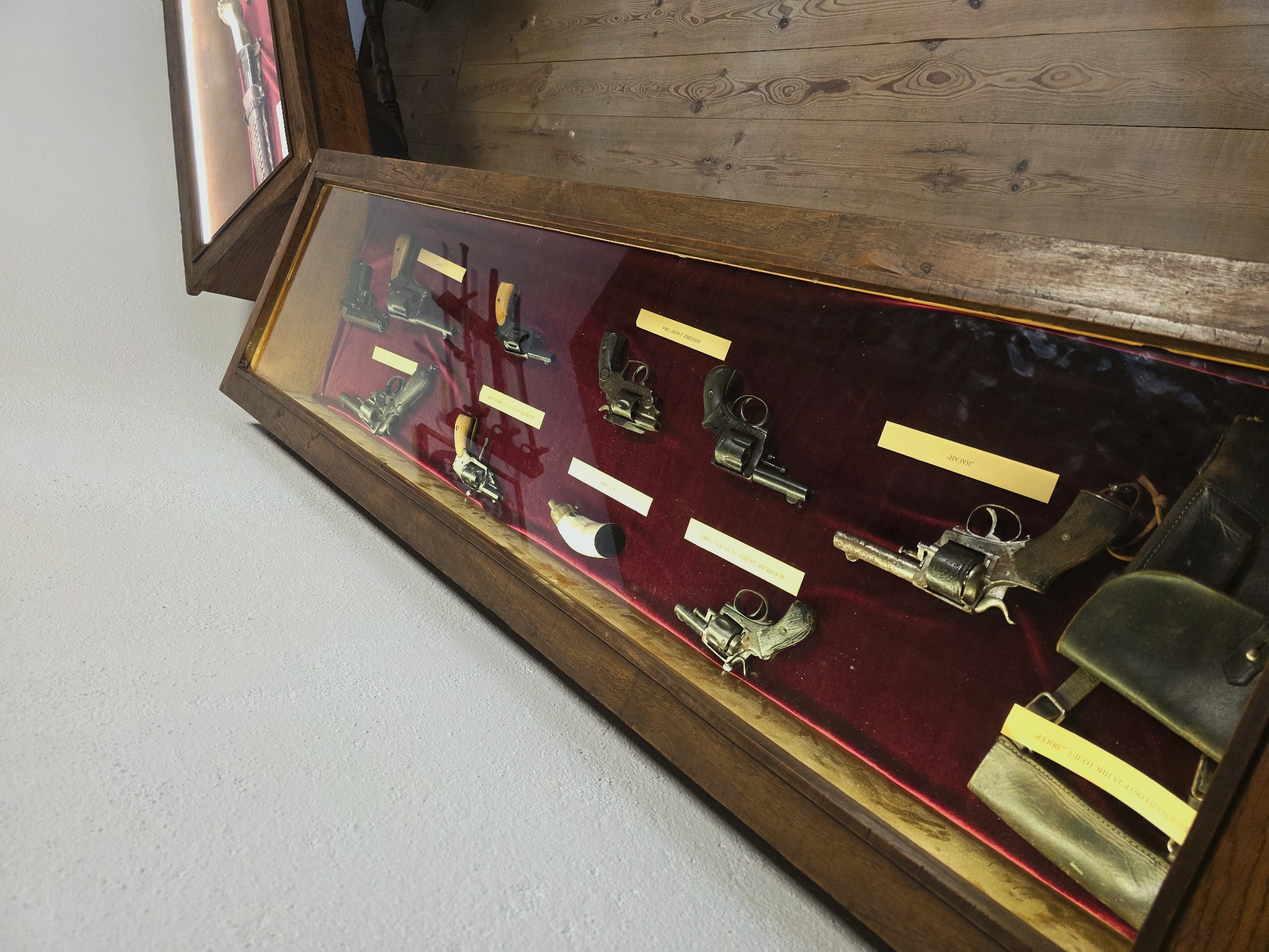 The image shows a museum display case containing historical firearms, specifically revolvers and possibly other handguns. The display case is made of wood with a glass front and is illuminated from within, highlighting the objects. The firearms are arranged on a deep red velvet lining, with several yellow descriptive labels placed in front of them. Some of the firearms appear to be old, with visible wear and patina. Two leather holsters are also present in the foreground of the display case. In the background, another display case with a sword and scabbard is partially visible. The floor is made of wooden planks. The overall setting suggests a museum or historical exhibition. The location is identified as Kovachevitsa, Bulgaria, implying this is a display within a museum or historical site in that town. No people are visible in the image.