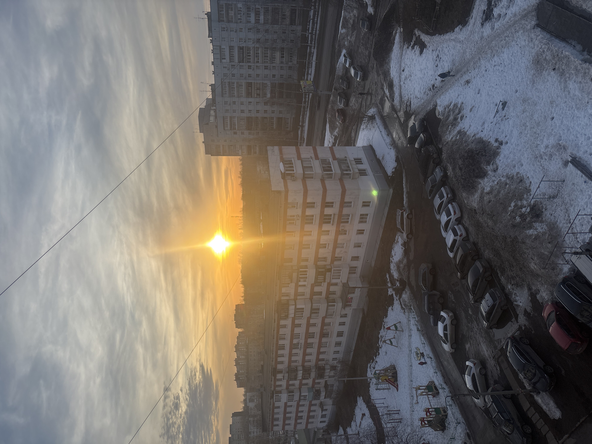 An elevated, outward-looking view captures a residential area in Chaikovsky, Russia, during what appears to be late winter or early spring at sunrise or sunset. The sun, a bright orb, is positioned in the upper-right quadrant, casting a strong golden glare across the scene and illuminating the lower sky with warm orange hues that transition to a soft blue-gray overhead. A distinct sun pillar is visible directly above the sun.
In the mid-ground and extending to the left, a five-story apartment building with a white facade and prominent red horizontal stripes between floors occupies a significant portion of the frame. It features numerous windows and several enclosed balconies. To the right, behind a road, a taller, multi-story (approximately nine to ten stories) gray apartment building stands, with numerous visible windows and roof-mounted antennae. Additional indistinct apartment buildings are visible in the hazy distance.
The ground level features widespread patches of dirty, melting snow, interspersed with bare ground, wet asphalt, and visible puddles. In the mid-left, two playground structures, partially covered in snow, are situated within a fenced area; one includes a slide and swings, the other appears to be a climbing frame. Numerous passenger vehicles, including sedans, SUVs, and hatchbacks of various colors (predominantly white, gray, and black), are parked along roads and in designated, snow-dusted parking areas.
Two individuals are discernible: one small figure, possibly a child, stands near the playground structures in the mid-left, and another person in dark clothing walks on a snow-covered path in the mid-right, moving away from the viewer. A third individual is standing near parked cars further down the street on the right. Overhead, a series of thin electrical or utility wires diagonally traverse the upper portion of the sky. Bare trees are visible in the background, particularly in the forested area beyond the buildings.
