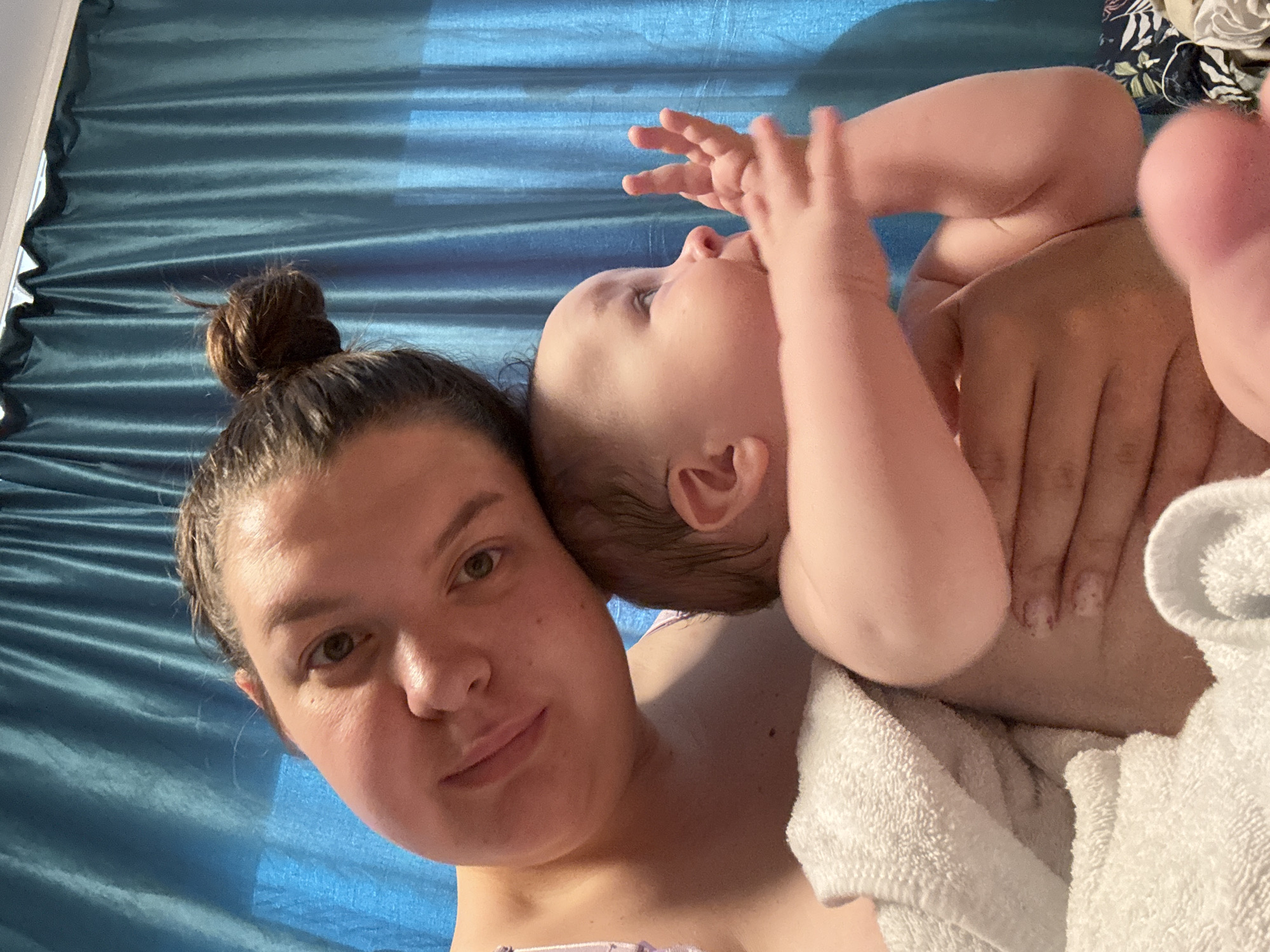 A woman with her hair in a bun is lying on a blue surface, possibly a bed. A baby is lying on top of her, with their hands raised near their face. The baby is partially wrapped in a white towel. The location is not discernible in the image, and no information is available about the city of Марковский, Russia.