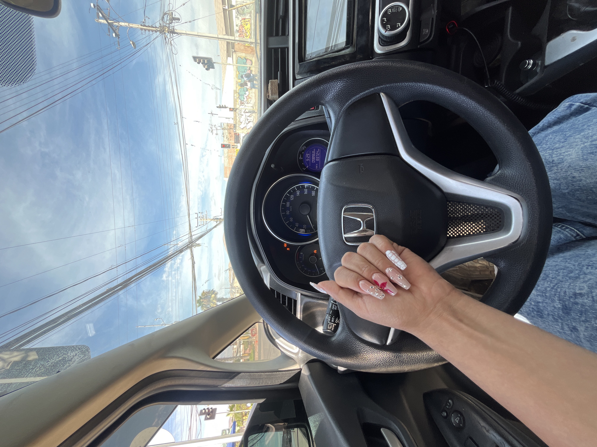 The image depicts the interior of a car, specifically the driver's perspective. A person's hand is visible on the steering wheel, adorned with decorative artificial nails. The car is a Honda based on the steering wheel emblem. The dashboard and speedometer are in focus. Outside the car, power lines, traffic lights, and buildings suggest an urban or suburban setting. Graffiti can be seen on the buildings. The location is Unknown, Unknown.