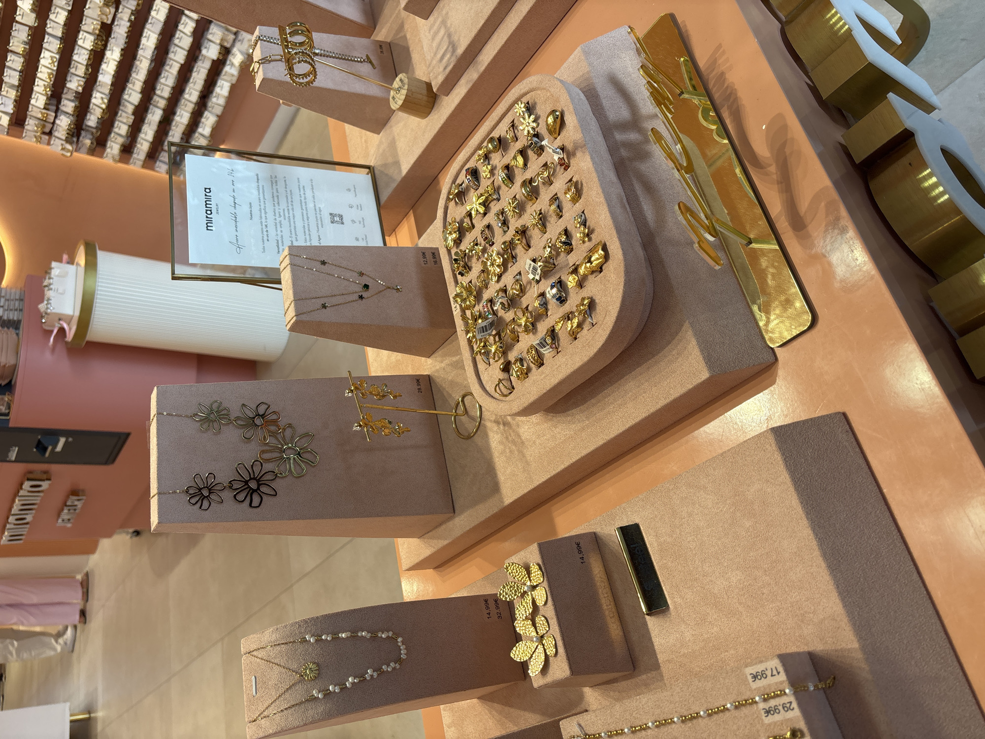 The image depicts a retail display of jewelry in a store. The jewelry includes necklaces, earrings, and rings, primarily made of gold-toned metal, with some pearl and green stone accents. The items are arranged on pink velvet display stands and a tray. In the background, there is a display wall covered with numerous pairs of earrings. A sign with the text "miramira JEWELRY" is visible on a stand. The overall setting appears to be a well-lit boutique with a modern and elegant aesthetic. The floor appears to be tiled. Two individuals are partially visible in the background, wearing pink pants and white tops, suggesting they are employees or customers. The image is taken from a medium-low angle, focusing on the jewelry displays. There is no information in the image to suggest the specific location within the city of Orihuela, Spain, other than it being a jewelry store.