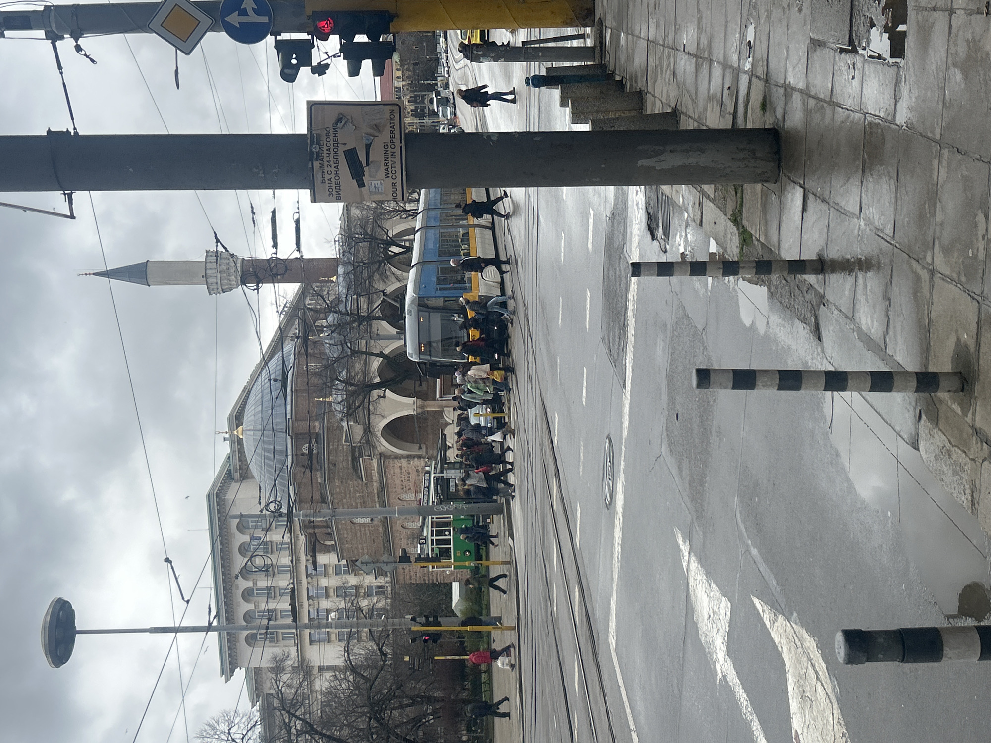 The image captures a rainy street scene in Sofia, Bulgaria. Pedestrians are crossing the street, near a blue and white tram. A green tram is also visible in the background, along with a building with a dome and arches. A minaret is visible. The street is wet and reflective. There are traffic lights and street signs on poles. The warning sign is in both Bulgarian and English: "WARNING! 24 HOUR CCTV IN OPERATION."