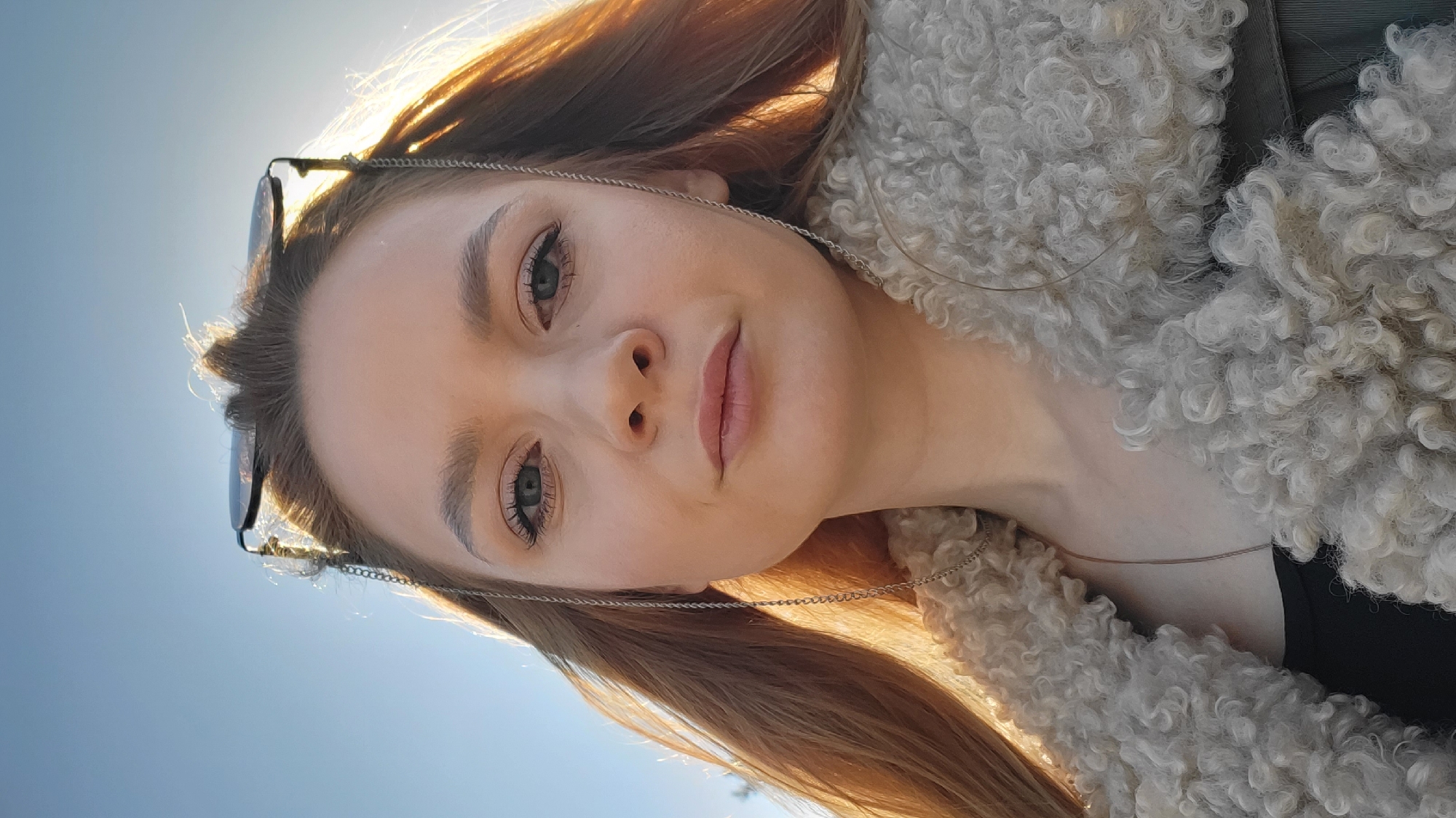A close-up, slightly rotated portrait shows a fair-skinned woman with reddish-blonde hair and light eyes, looking directly at the camera. Sunglasses are perched on top of her head, attached by a silver chain. She wears a light-colored, textured, possibly fleece or faux fur jacket. The background reveals a clear, light blue sky, suggesting an outdoor setting. The subject is likely located outdoors. There is no indication of a location in the city of Chaikovsky, Russia.