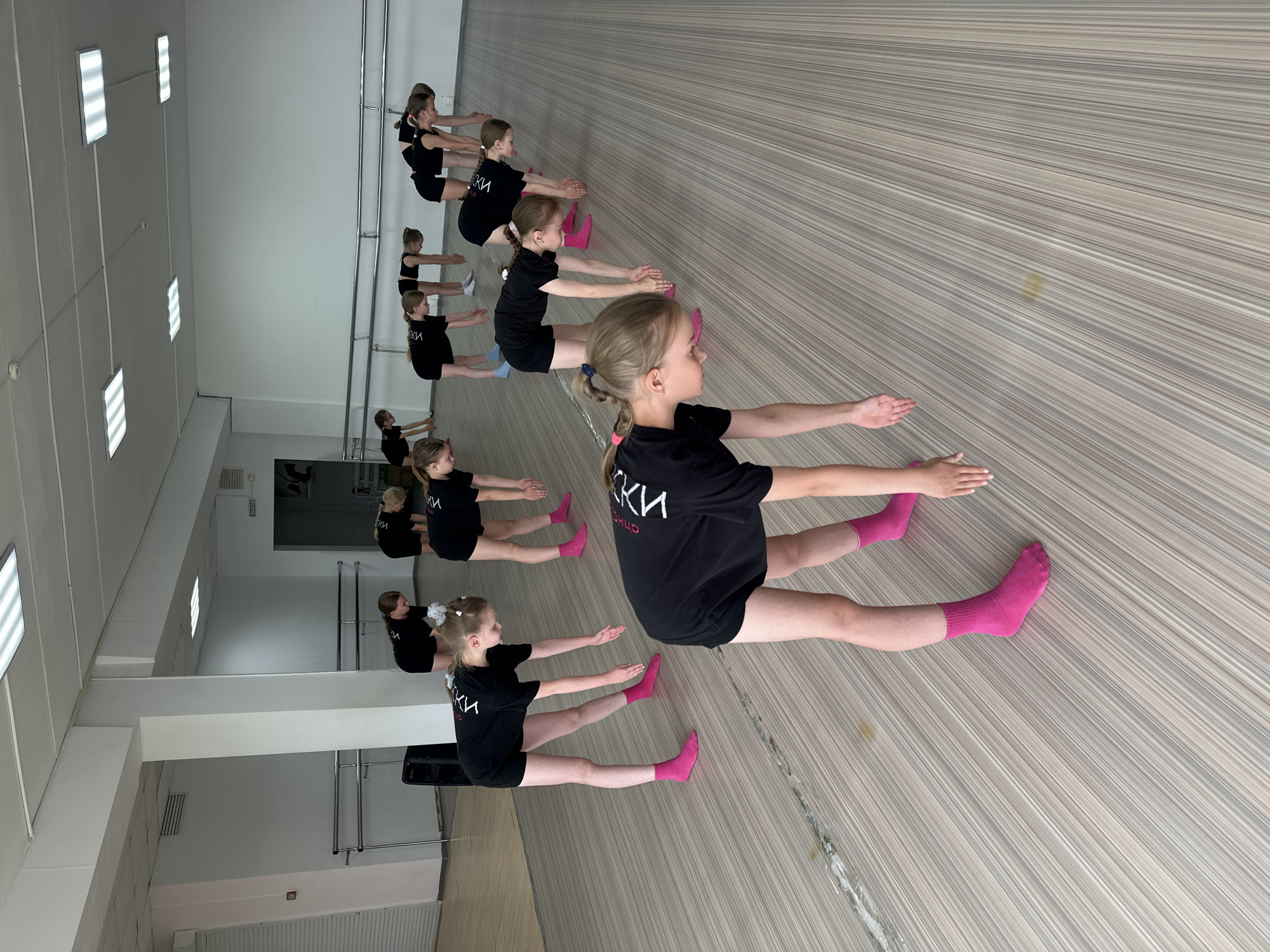 A ballet class takes place in Votkinsk, Russia. Approximately 10 young girls, all wearing black t-shirts with white lettering (possibly Cyrillic) and shorts, are stretching on the floor in a line. Most are wearing bright pink socks. They are seated with their legs outstretched and reaching their arms forward. The room has a light-colored, horizontally lined floor, a white wall, and a barre is visible in the background along the wall. Overhead are rectangular ceiling lights. A closed door and air conditioning vent are seen on the left side of the image.