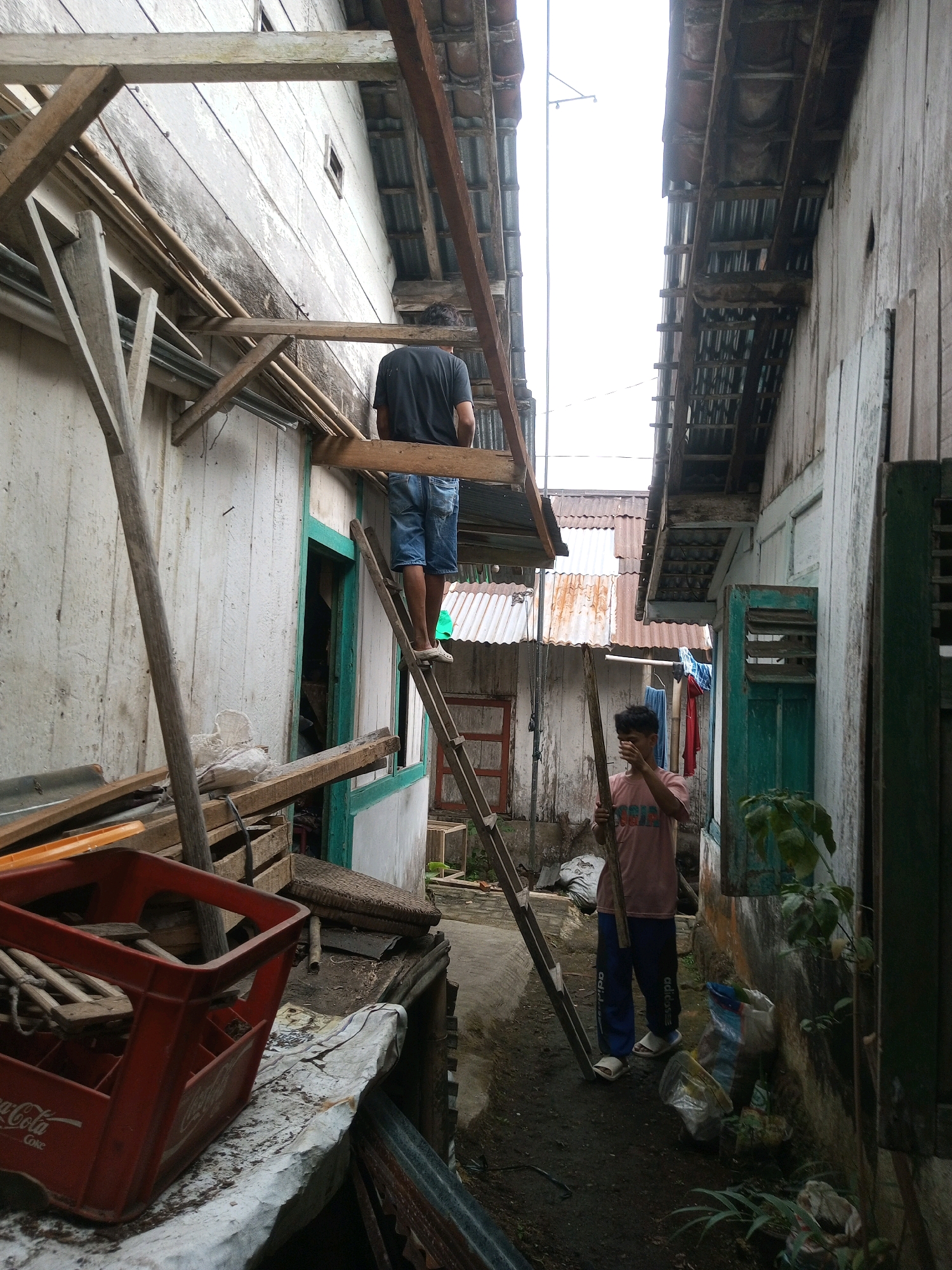 A young man in blue track pants with white lettering and a pink t-shirt stands on a dirt path, holding a wooden pole horizontally in front of him, partially obscuring his face. He is positioned between two weathered, light-colored buildings. To his left, a wooden ladder leans against the side of the building, with another person wearing a black t-shirt and denim shorts standing on the third rung. The buildings are constructed with overlapping wooden planks and corrugated metal roofing. The left building has a green-trimmed window. Debris, including lumber, a red plastic crate, and plastic bags, is scattered on the ground and on a ledge in the foreground. Clothes hang on a line between the buildings in the background. The scene appears to be an alleyway in Andongsili, Indonesia, possibly undergoing construction or maintenance.