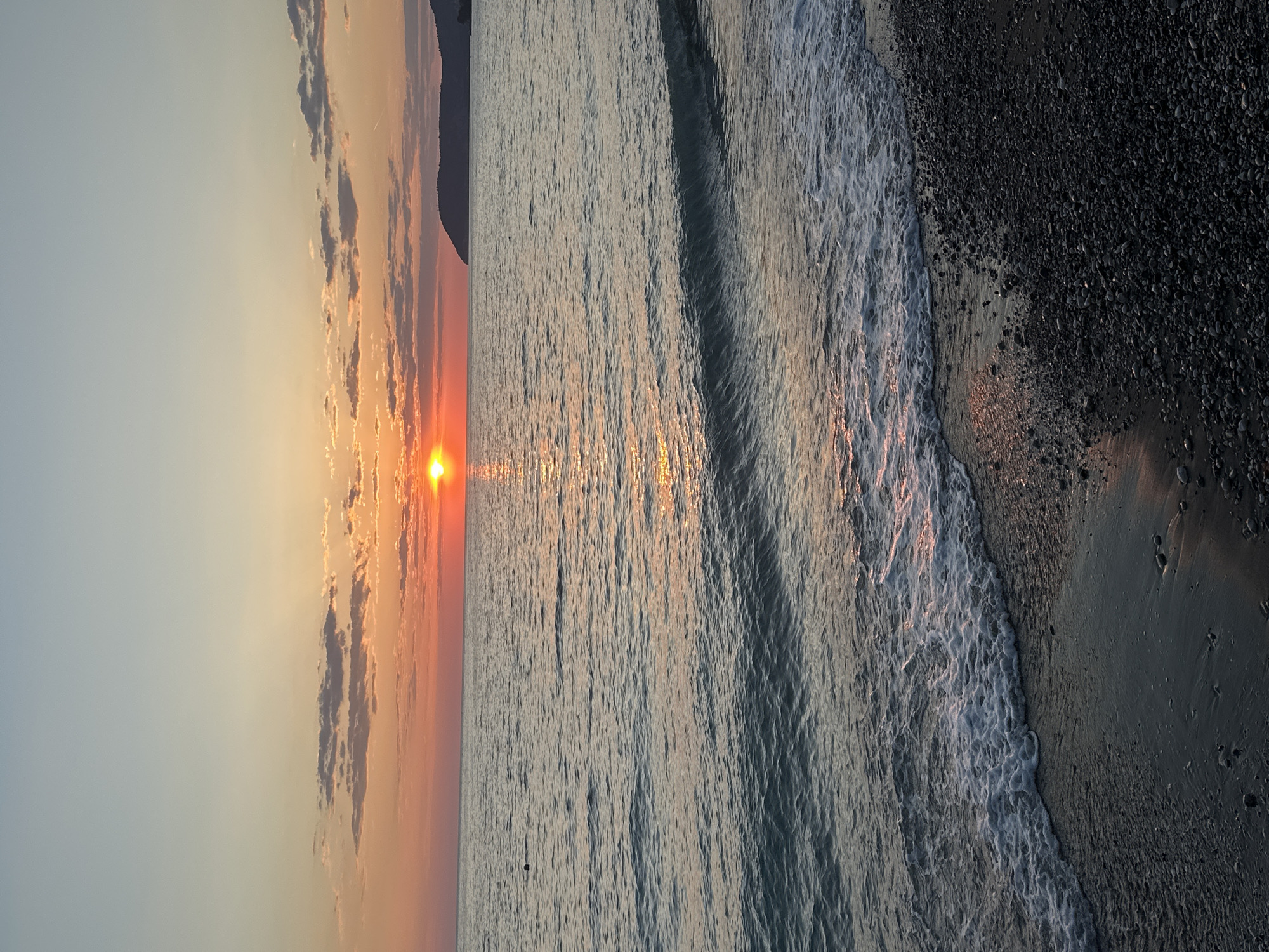 The image shows a sunset over the Adriatic Sea in Šušanj, Montenegro. The sun is setting on the left side of the frame, casting an orange glow on the sky and water. The sea is calm, with small waves washing onto a dark, rocky beach. The beach occupies the right side of the frame. There are no people visible in the image. A mountain can be faintly discerned on the far left side of the frame.