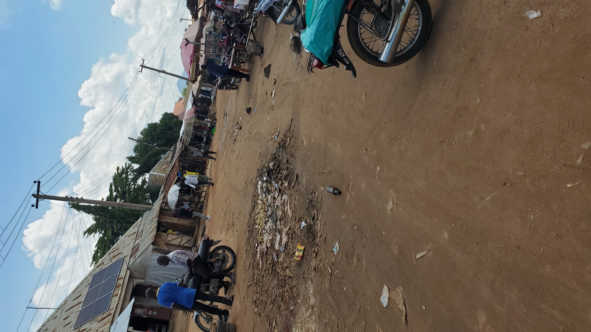 A dusty, outdoor marketplace in Jalingo, Nigeria, is depicted under a partly cloudy sky. The scene is dominated by dirt ground and several market stalls with corrugated metal roofs. On the left, a building with solar panels is visible. Several individuals are present, some standing near motorcycles, while others appear to be interacting at the market stalls. A pile of debris, including dried leaves and scattered trash, is prominent in the foreground. Multiple motorcycles are parked throughout the scene, some with riders, and one is partially covered with a green tarp. Electrical poles and wires crisscross the sky. The overall impression is of a busy, functional marketplace with elements of everyday life.