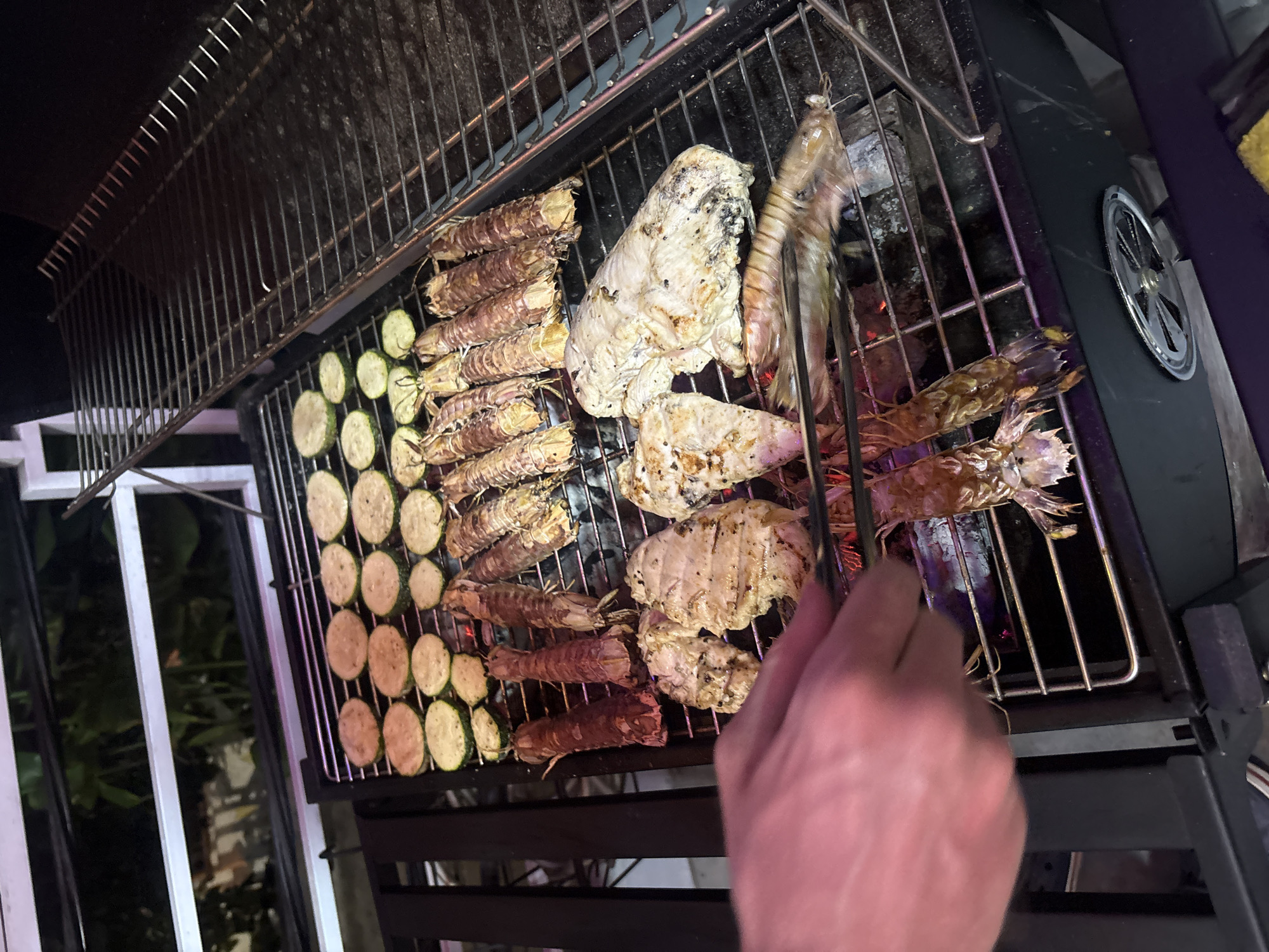 In Nha Trang Ward, Vietnam, a grill laden with food is visible. The grill contains sliced zucchini, several mantis shrimps, and cuts of chicken. A hand holds tongs and manipulates the food on the grill. The grill appears to be outdoors, with a window and possibly some vegetation visible in the background. The lighting is dim, suggesting it could be evening.
