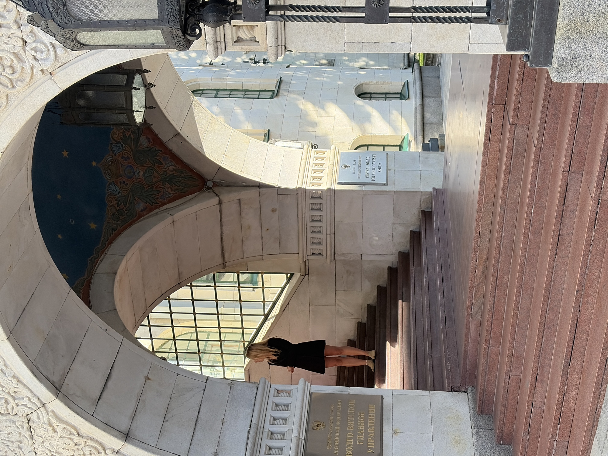 The image captures a woman standing on a staircase in front of a stone building with arched entrances. The architecture features ornate carvings and a painted ceiling with stars. A sign on the building reads "CENTRAL BANK OF RUSSIAN FEDERATION CENTRAL BOARD FOR VOLGO-VYATSKY REGION". The woman is wearing a black dress and light-colored shoes and is looking to her left. The scene is illuminated by sunlight, casting shadows on the building and steps. This building is the Volgo-Vyatka Main Branch of the Bank of Russia, located in Nizhny Novgorod, Russia.