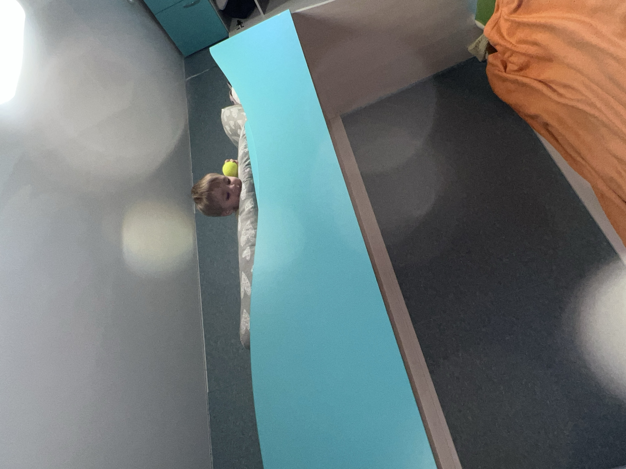 A young child is lying on a bed next to a teal-colored table or cabinet. The child is holding a yellow ball. The bed is covered with a patterned blanket. An orange blanket is visible in the background. The location of Noviy, Russia, is not possible to verify based on the image alone.