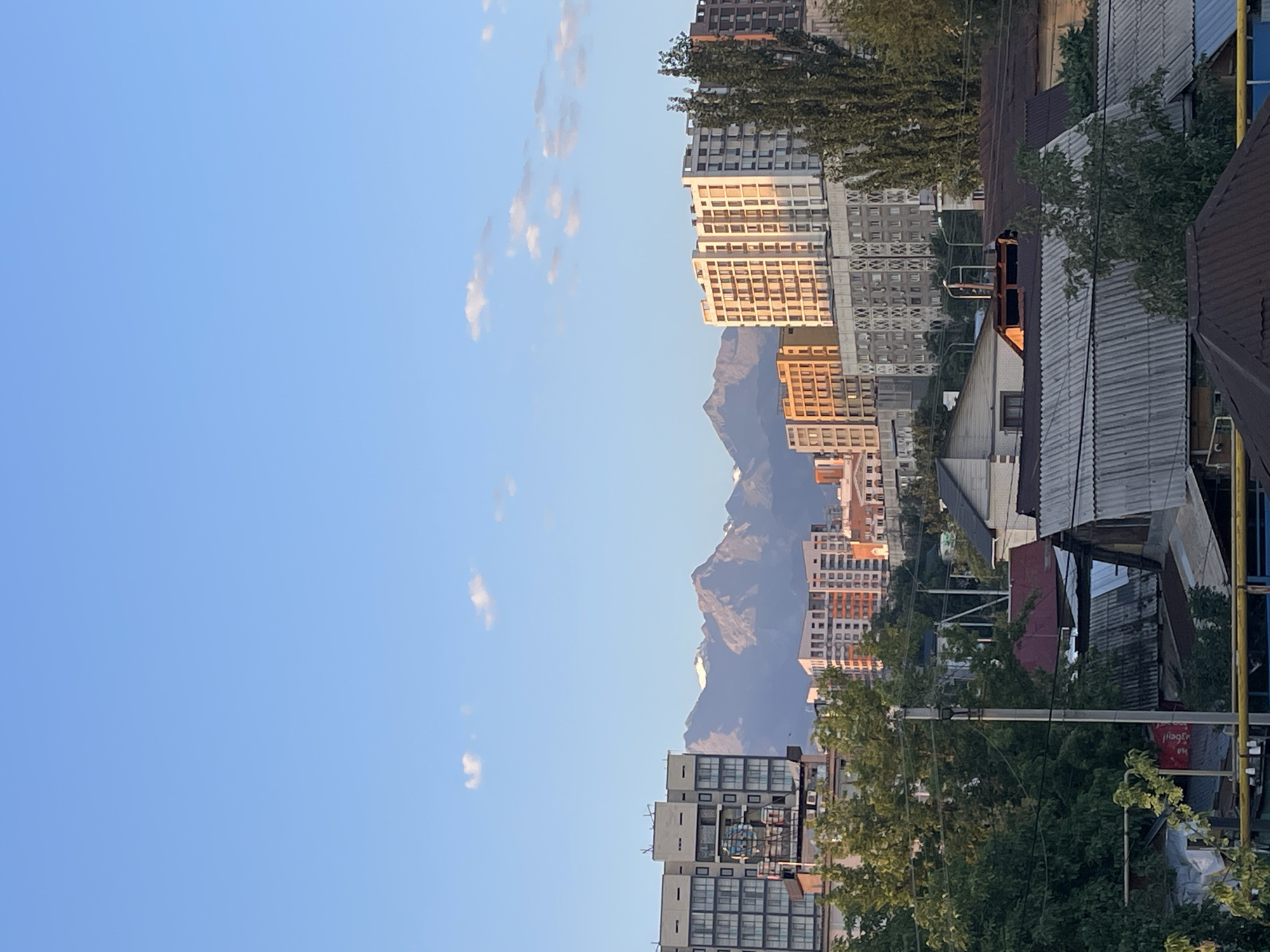 This is a vertical, wide-angle photograph capturing an urban landscape in Almaty, Kazakhstan, with mountains in the background. The scene is illuminated by the warm glow of what appears to be late afternoon or early morning sun.
In the foreground, the tops of several low-rise buildings with corrugated metal roofs are visible. These structures are primarily clad in muted tones of gray and brown, suggesting they may be older residential or utility buildings. Some of these rooftops feature visible piping and what appears to be ventilation systems. Trees with dense green foliage are interspersed among these buildings, some reaching up to obscure parts of the mid-ground.
The mid-ground is dominated by a collection of multi-story apartment buildings. To the left, a tall, modern building with a facade of glass and dark panels stands prominently. Next to it, several other apartment blocks are visible, featuring a mix of architectural styles and colors, including light beige, orange, and white. Some of these buildings exhibit a grid-like pattern of windows. The sunlight highlights the facades of some of these structures, creating a dappled effect. Tall, slender trees with dark green foliage are also present in this section of the city.
The background is marked by a majestic mountain range, possibly part of the Tian Shan mountains that surround Almaty. The peaks are jagged and appear to be snow-capped, shrouded in a light haze that softens their outline against the clear blue sky. The sky itself is a vibrant, cloudless blue, with a few wispy white clouds scattered across the upper portion of the frame.
There are no people or animals visible in the photograph, and no overt actions or interactions are depicted. The environment is clearly urban, with a mix of residential and commercial architecture, set against a dramatic natural backdrop. The overall impression is one of a city integrated with its mountainous surroundings.