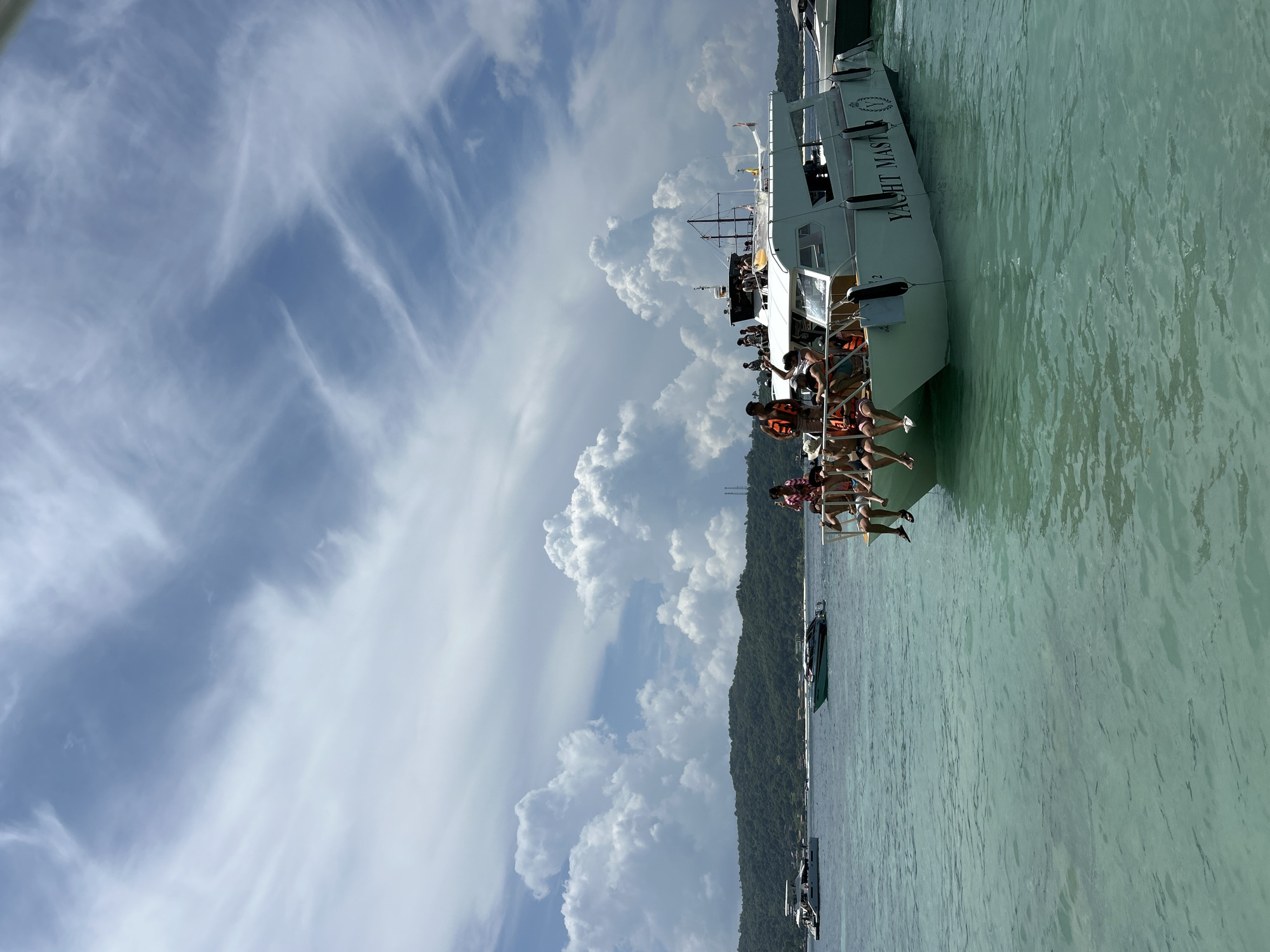 The image depicts a sunny day on the turquoise waters near Ko, Thailand. A white motor yacht named "YACHT MASTER" is prominently featured, with several passengers seated along its bow, many wearing life vests. The boat is on the water, which appears shallow enough to see the sandy bottom in some areas. In the background, lush green islands rise from the sea, covered in dense vegetation. Other boats, including a smaller speed boat, are also visible in the distance. The sky is a bright blue, adorned with scattered white cumulus clouds. The overall scene suggests a leisure outing or a tour.