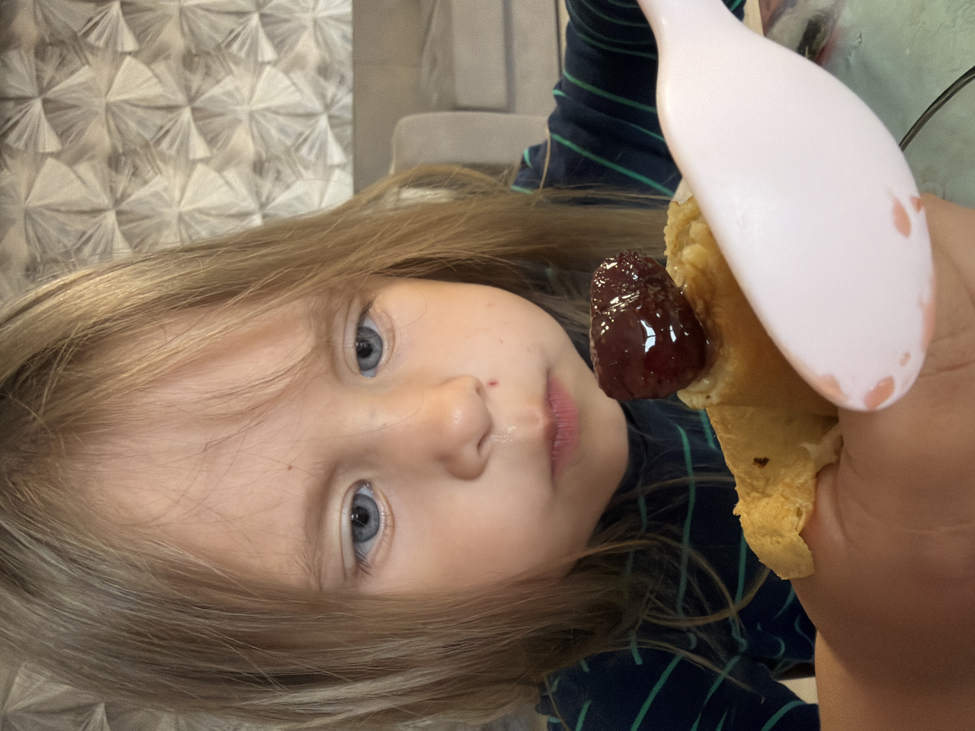 This close-up, rotated portrait captures a young child with long, light-colored hair and distinct blue eyes looking directly at the viewer. Her face fills the left side of the frame, with a neutral and slightly curious expression, and her lips are slightly parted. She is dressed in a dark blue garment with green stripes.
In the foreground on the right, the child holds a piece of a light-colored pancake or crepe, which is topped with a glistening dollop of dark red jam or fruit preserve. A light pink spoon, held by the child's hand, is positioned near the food, indicating an ongoing meal. The spoon also has some reddish residue on it.
The background behind the child's head features a patterned wall or wallpaper in light grey or beige, displaying a repetitive geometric, fan-like design. The scene is set indoors. No specific time of day or weather is evident, and there is no visible text. The location context for this activity is Chaikovsky, Russia.