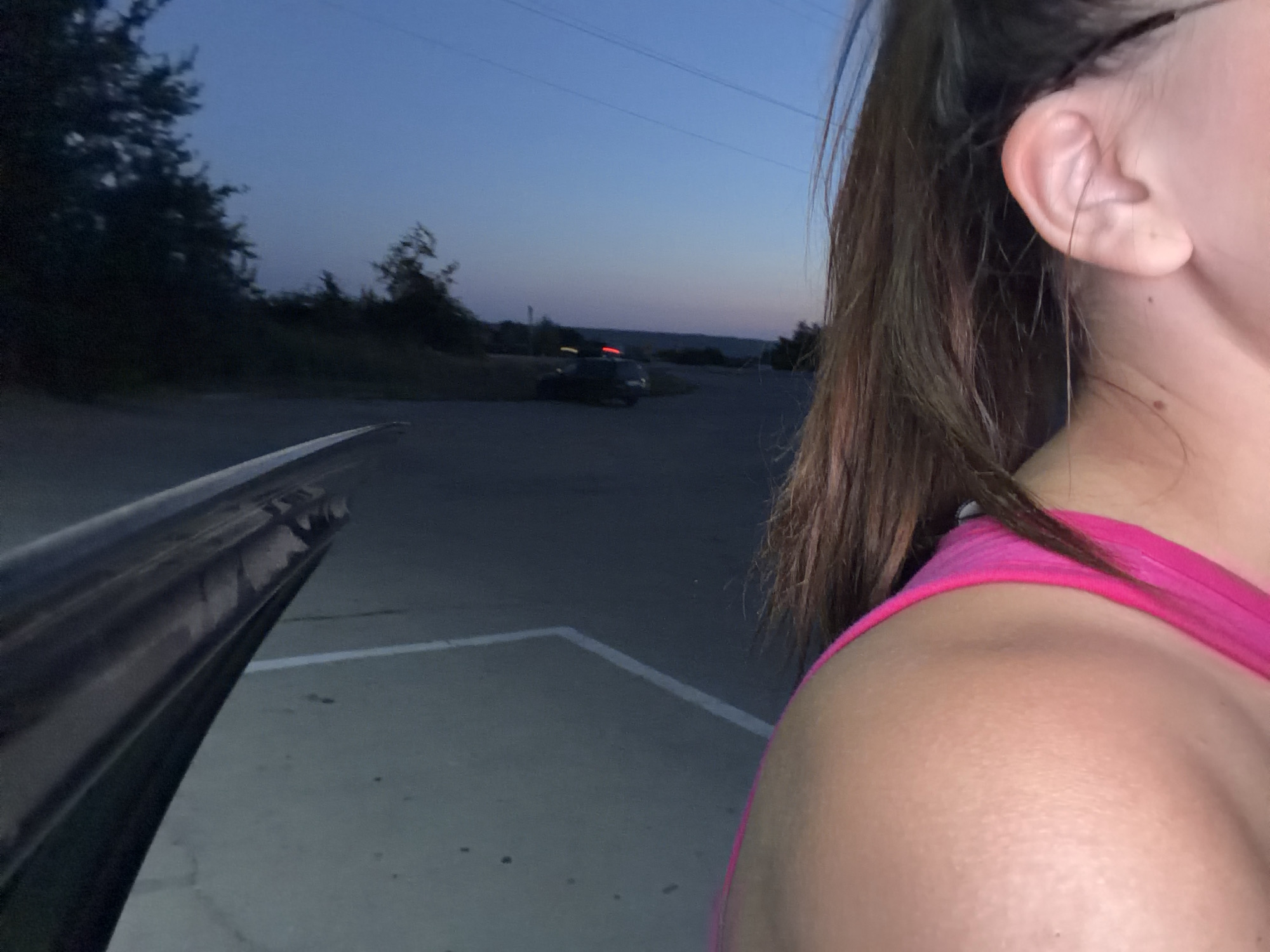 The image depicts a close-up, low-angle shot of a woman's ear and shoulder, with her right side dominating the frame. She is wearing a bright pink tank top. Her dark hair is pulled back in a ponytail. The background suggests an outdoor setting at dusk, with a twilight sky and silhouetted trees. A dark-colored car is visible in the distance, parked on a paved surface marked with white lines. A metal object, possibly part of a vehicle or structure, is in the foreground on the left. The scene does not provide any specific information to identify the location as Orhei, Moldova, or any other particular city.