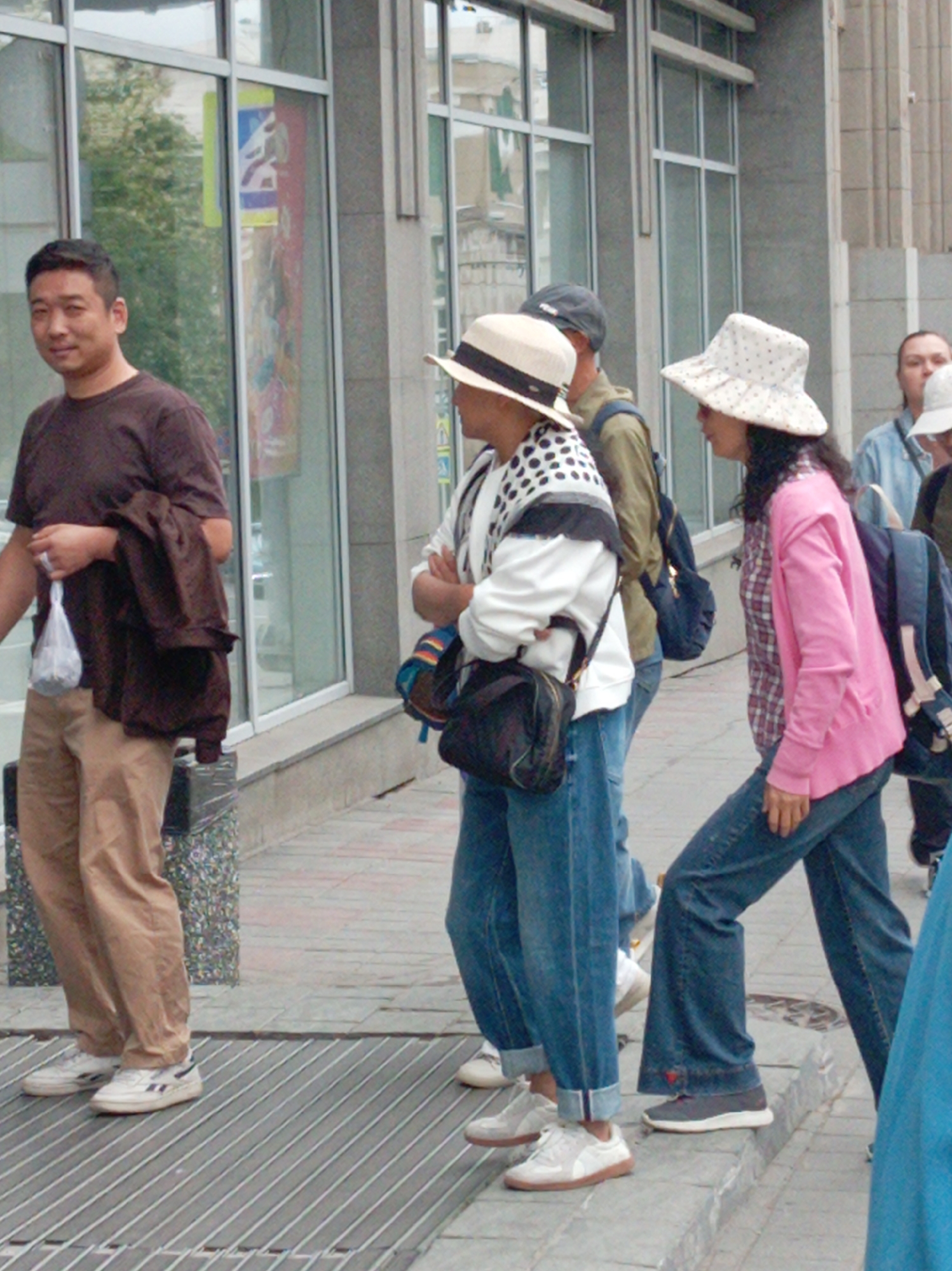 A group of people are walking on a city sidewalk in Vladivostok, Russia, near a building with large windows. A man in a dark brown shirt and tan pants is in the foreground, holding a plastic bag and a dark jacket over his arm. He is looking towards the left. Next to him, a woman wearing a white patterned top and jeans, and a straw hat, is walking away from the camera. Behind them, another woman in a pink cardigan and jeans, wearing a white polka-dot hat, is also walking away. Further back, a man wearing a blue shirt and a backpack is visible. The sidewalk is paved with brick and metal grating. The building has large reflective windows that show trees and the sky. The overall scene depicts a casual urban street setting.