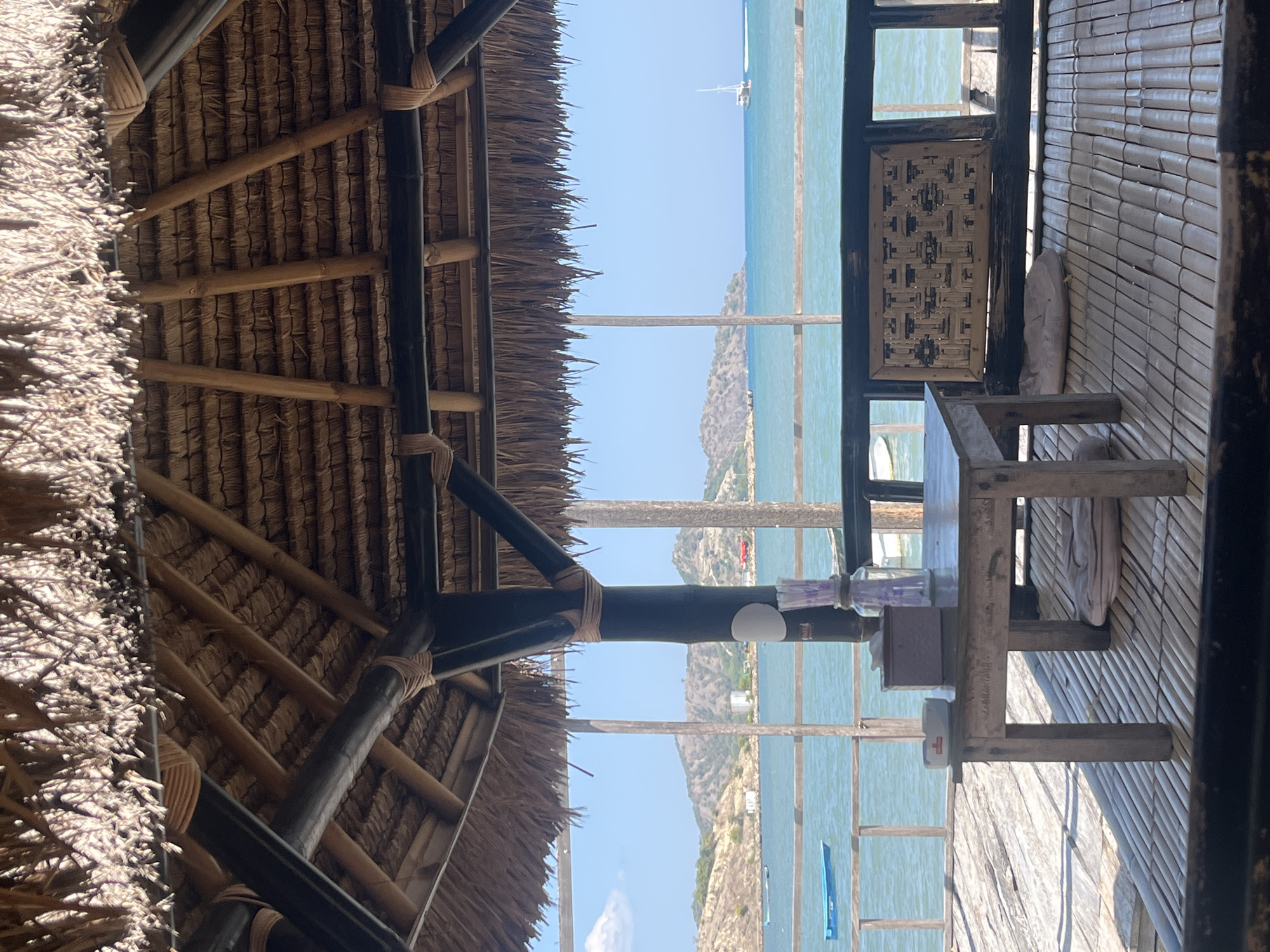 The image shows a view from under a thatched-roof gazebo, looking out over the ocean. In the foreground, there is a rustic wooden table and a bamboo floor. On the table, there is an ashtray, a box of tissues, and a glass jar filled with straws. In the middle ground, there is a railing overlooking the water, with a wooden bench on the right. The water is a clear turquoise color and there are several boats visible, including a small blue boat in the distance and a sailboat further out at sea. In the background, there are hills and mountains. The overall scene appears to be a peaceful and relaxing beachfront setting.