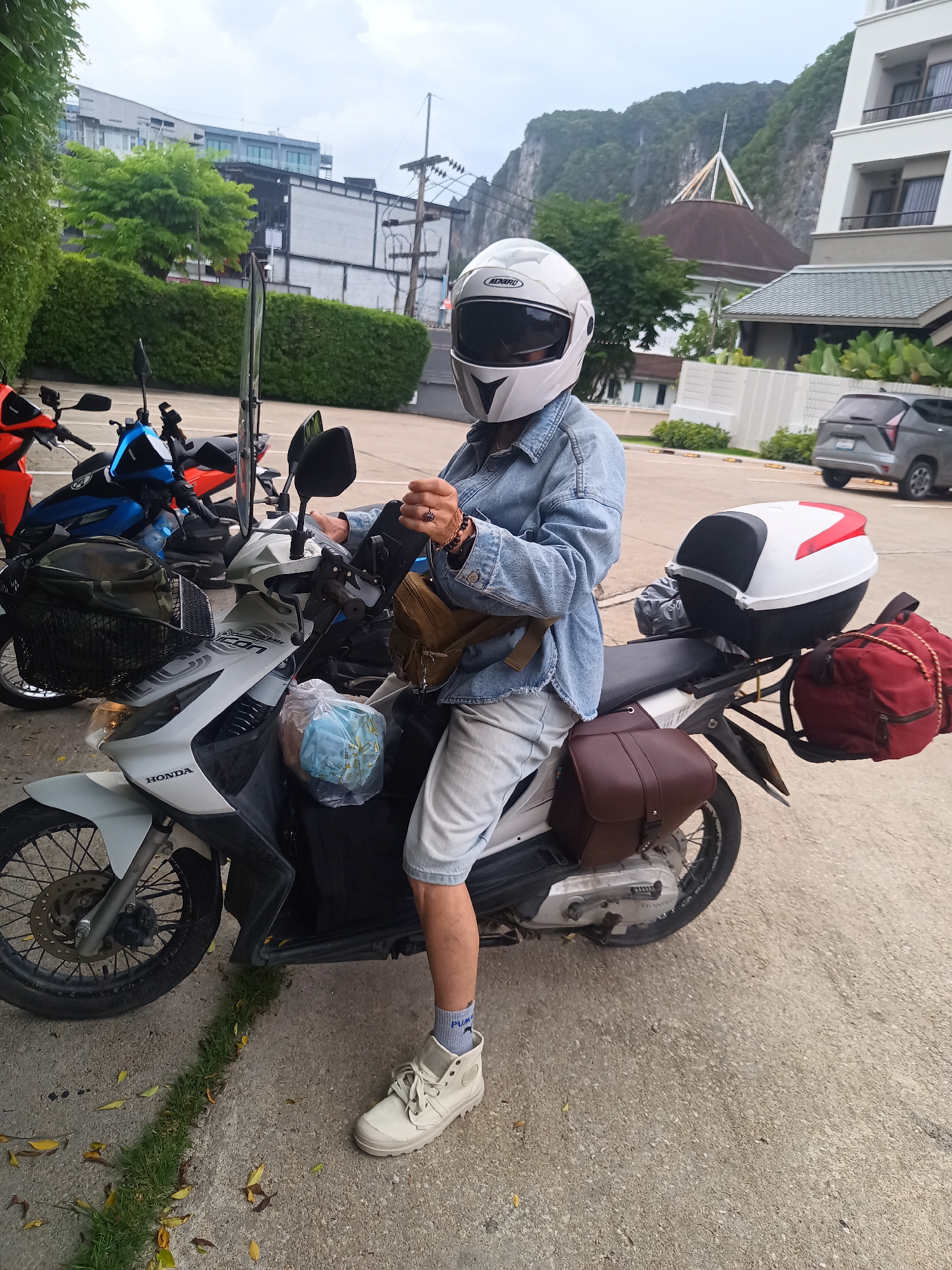 A person wearing a white helmet, denim jacket, and light-colored shorts sits on a white Honda motorcycle loaded with luggage, including saddlebags, a top box, and a red duffel bag, in what appears to be a parking area in Ban Ao Nang, Thailand. Other motorcycles are parked nearby. The background features buildings, trees, and a limestone karst mountain.