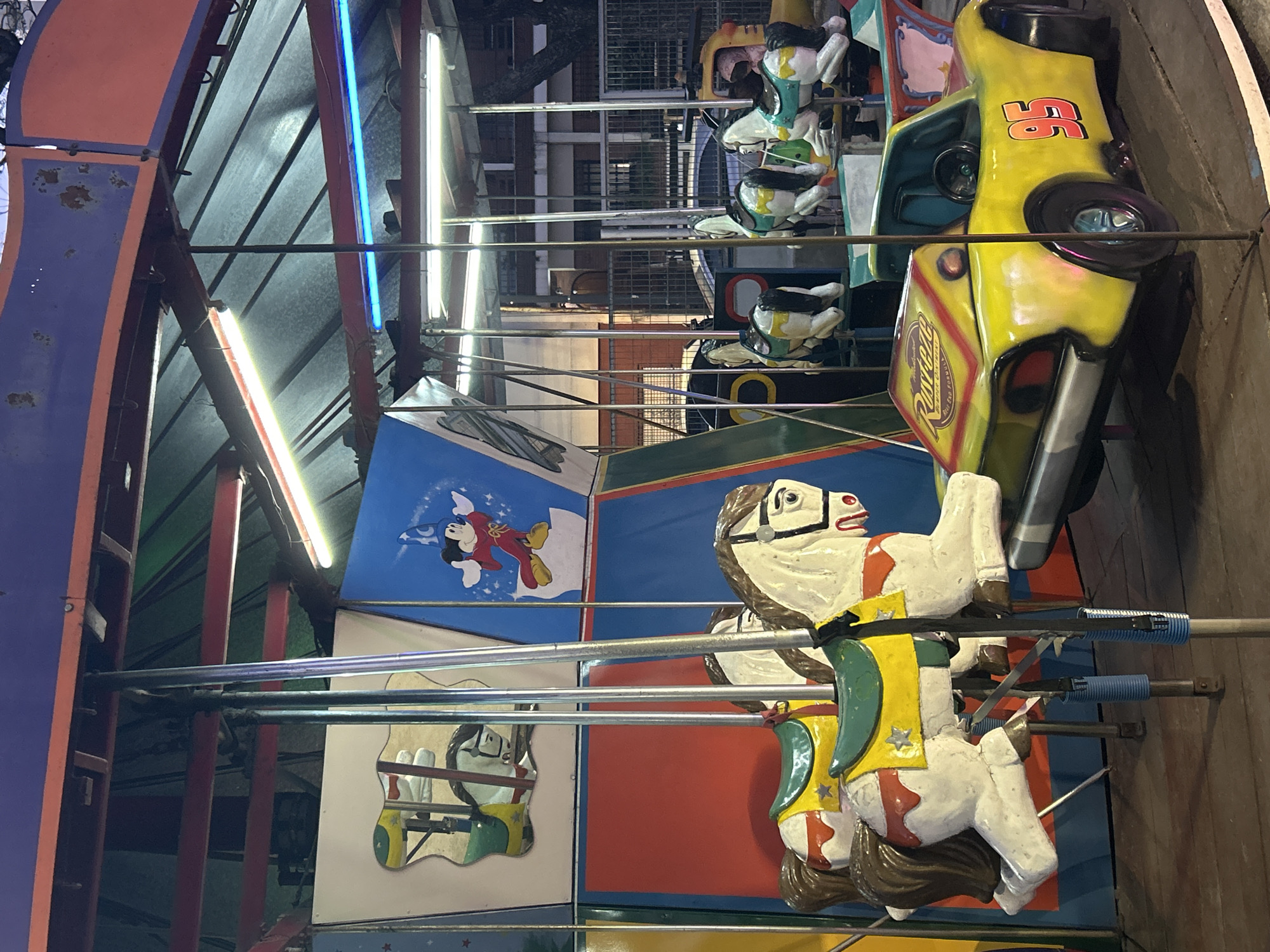 The image depicts a carousel ride in what appears to be an amusement park or fairground, possibly located in the city of Buenos Aires, Argentina. The carousel features several colorful amusement vehicles, including white horses with brown manes and tails, adorned with yellow and green saddles, and a yellow race car with the number "95" on its side. The race car has a "Rust-eze" logo on its rear. In the background, there are painted panels depicting cartoon characters, including Mickey Mouse. The structure of the carousel is visible, with metal poles, a blue and orange roof, and fluorescent lighting. The overall scene suggests a nighttime setting, with artificial lights illuminating the ride. Buildings and trees are faintly visible in the background, hinting at an urban environment. No people are clearly visible in the image, and the carousel appears to be stationary.
