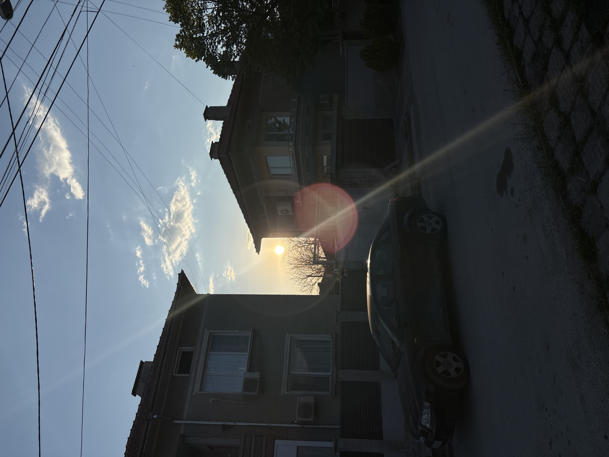 The image depicts a street scene in the city of Chirpan, Bulgaria, during late afternoon or early evening, indicated by the low sun. Two buildings of grey stone are visible, with a dark-colored car parked on the adjacent street. Numerous power lines are seen extending into the upper left. The street is relatively dark with a possible grass-covered paving.