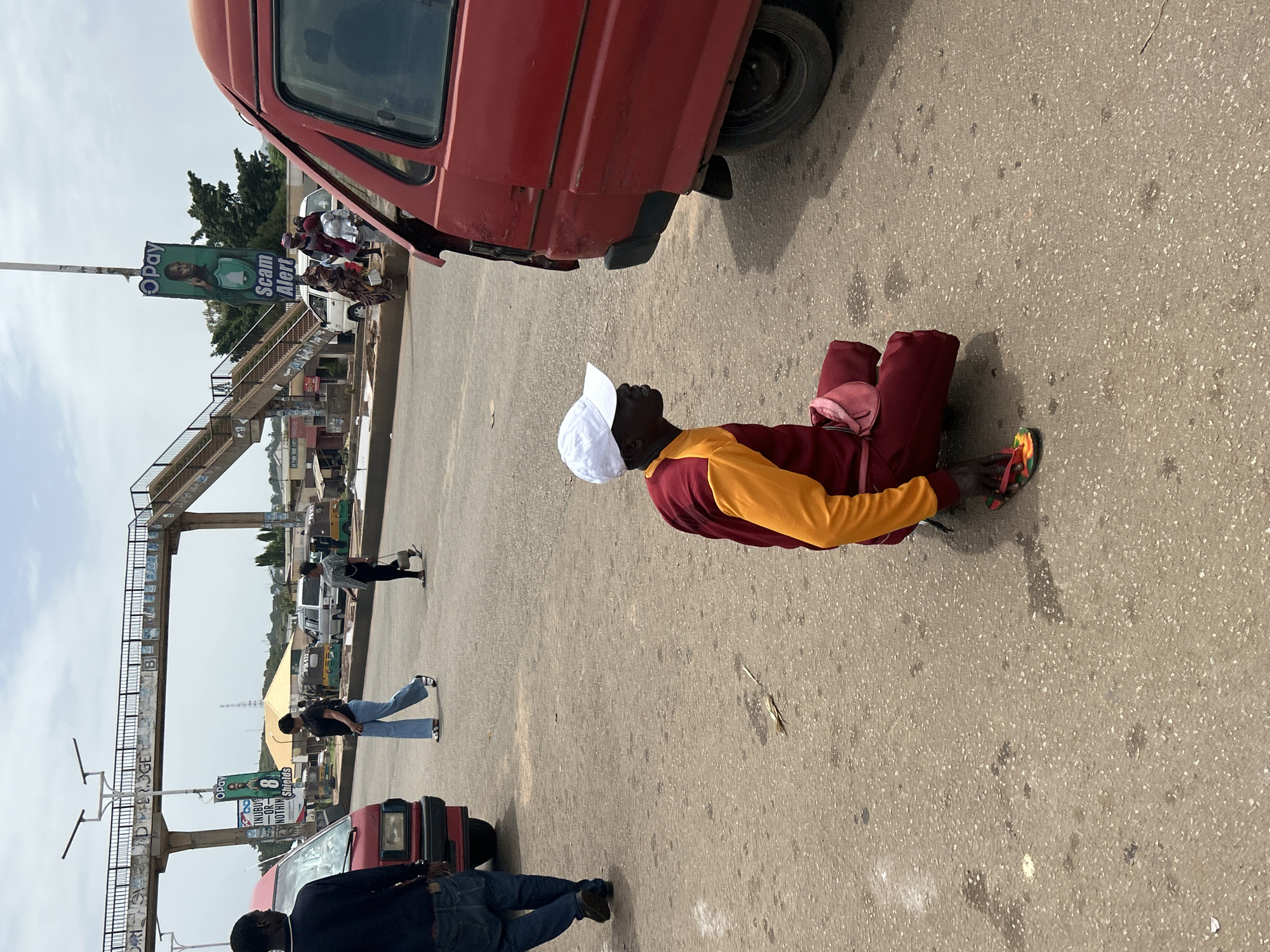 The image depicts a street scene in Jos, Nigeria. In the foreground, a person wearing a white baseball cap, a maroon and yellow long-sleeved shirt, and maroon pants is crouching on the asphalt. A small pink pouch is attached to their belt. They are wearing colorful flip-flops. To the right, the rear section of a red van is visible. In the background, a pedestrian bridge spans across the road, with various vehicles including a red van, a white mini-bus, and a few tricycles (keke napep) on the road. People are walking on the road and sidewalk. An advertising billboard for "OPay" with a "Scam Alert" message is prominently displayed on a pole. The sky is overcast.
