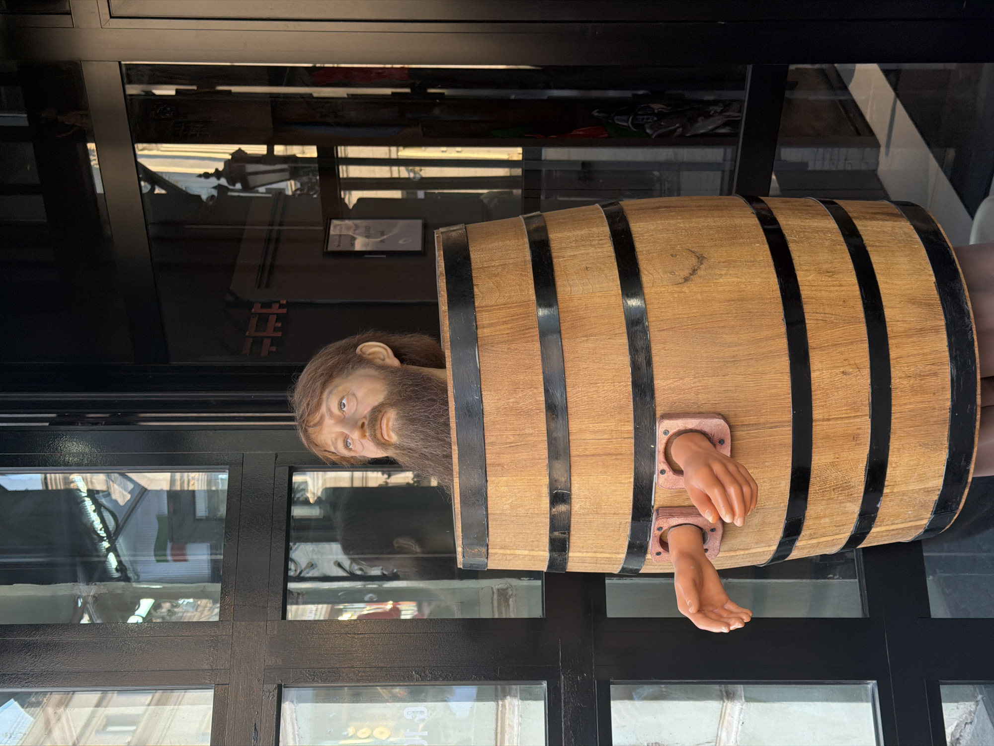 A mannequin depicting a person in a wooden barrel with restraints on the wrists is displayed in Kazan, Russia. The mannequin has a head and arms protruding from the barrel. The barrel itself has a wooden surface and is reinforced with dark metal bands. The scene is positioned outside a building with dark-framed glass windows and panels. The reflections in the glass indicate an urban setting.
